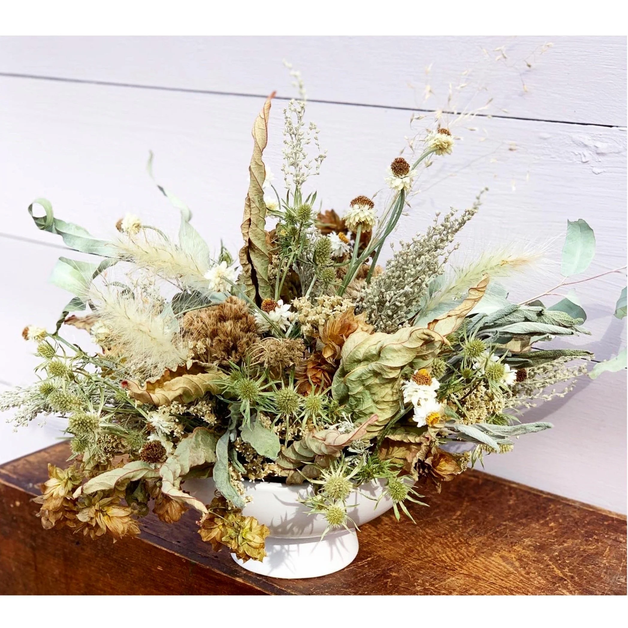 Dried flower arrangement in a white bowl, featuring various plants and branches with brown and green tones, placed on a wooden surface.