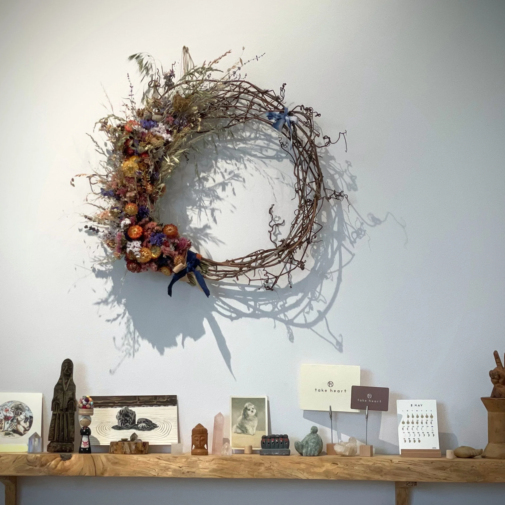 A decorative wreath made of intertwined branches with dried flowers and ribbons hanging on a white wall above a wooden shelf with various small decorative items and photographs.