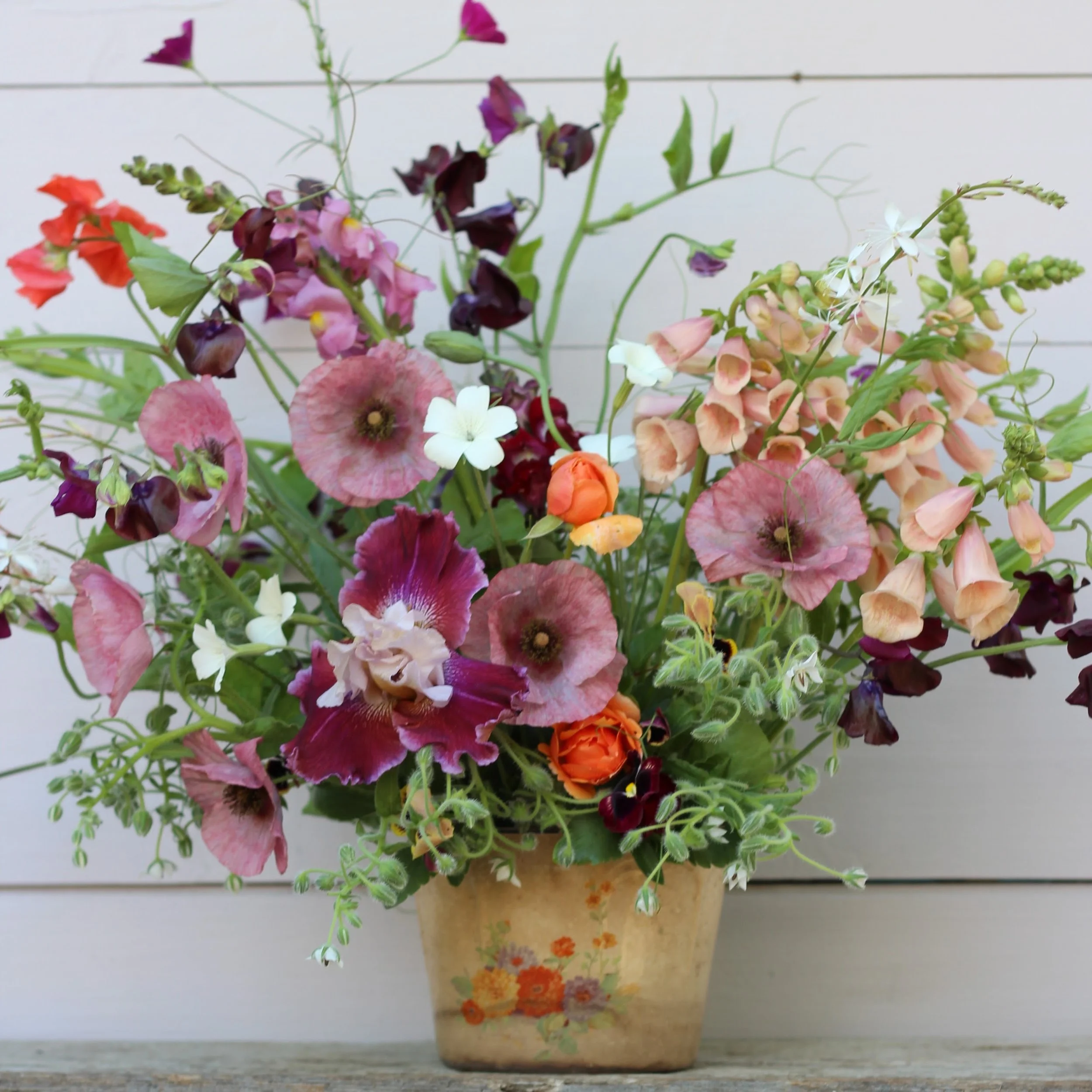 Colorful bouquet of various flowers in a painted beige pot with orange and yellow flowers, set against a white wooden background.