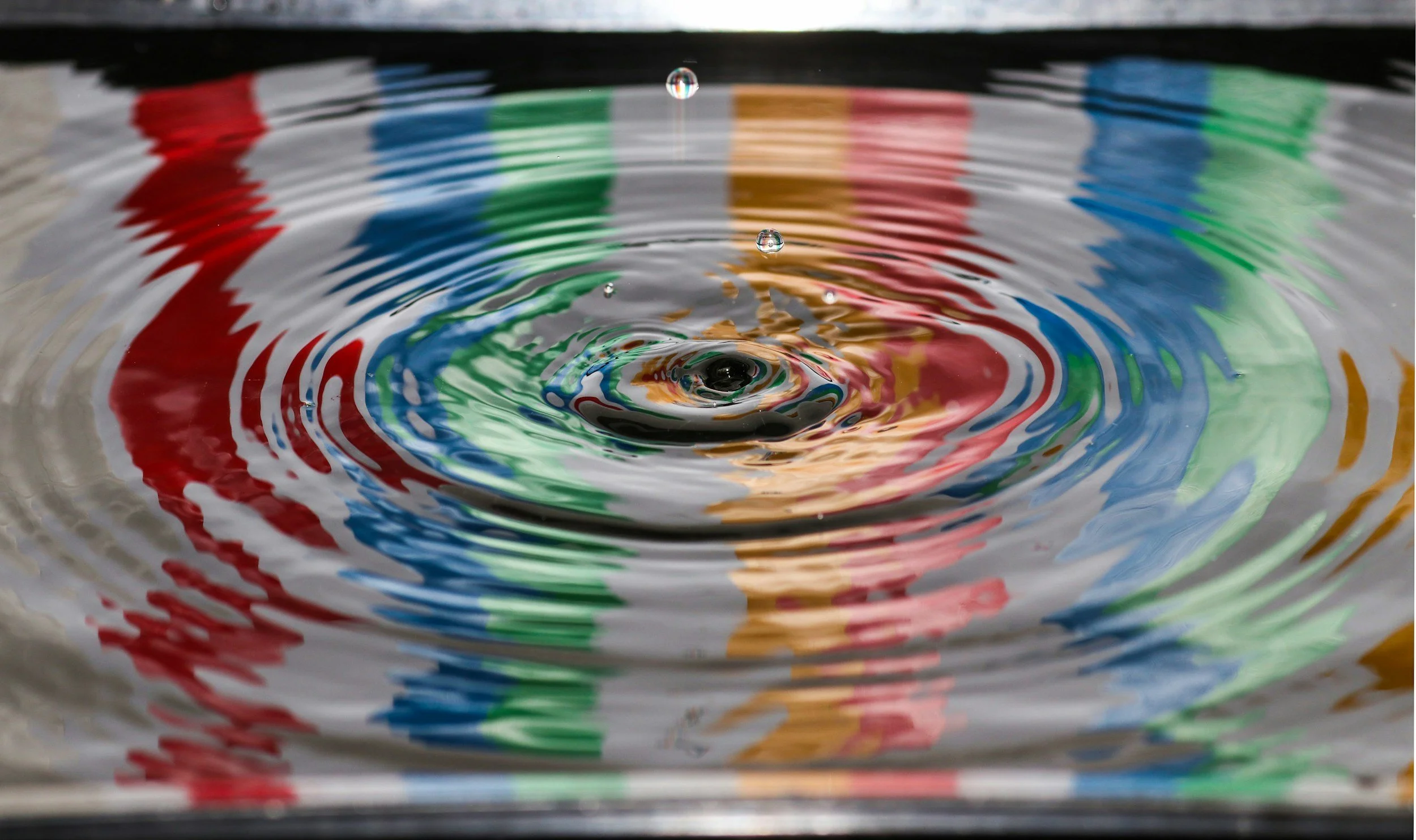 A droplet of water ripples outwards, the reflection shows colourful stripes