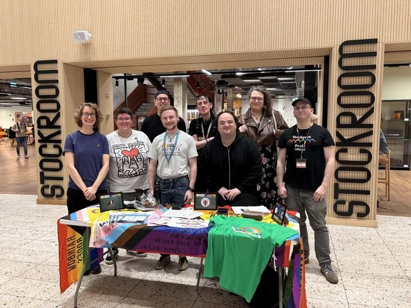 Image shows a group of diverse people stood around a table with rainbow banners on, in front of Stockroom