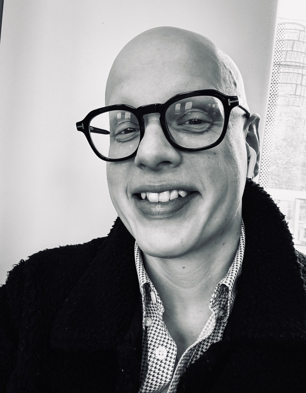 Black and white photo of a person with glasses, smiling, with a bald head and wearing a dark shirt.