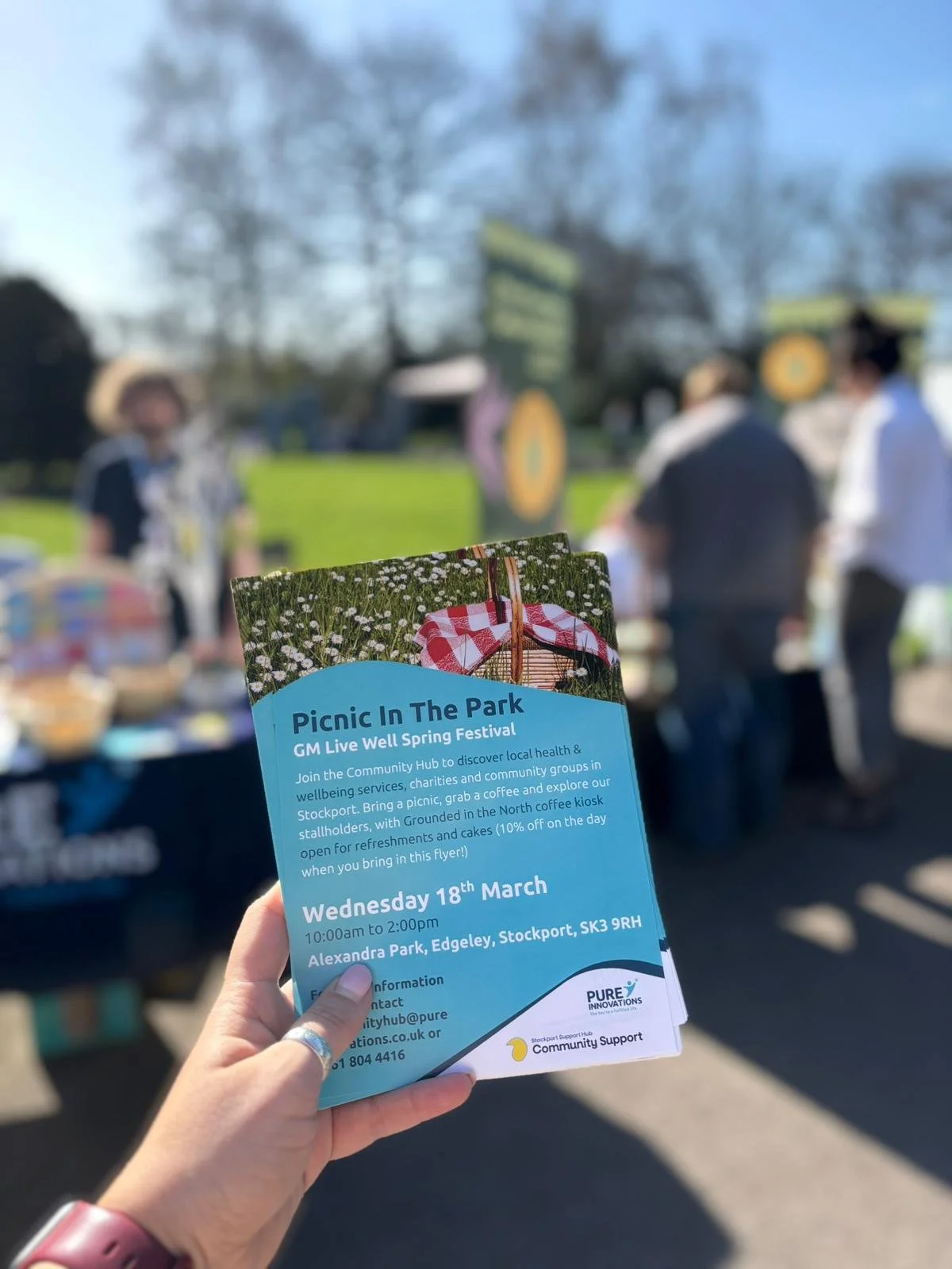 Image shows a close up of a 'Picnic in the Park' flyer, in the background people are gathering around various stalls