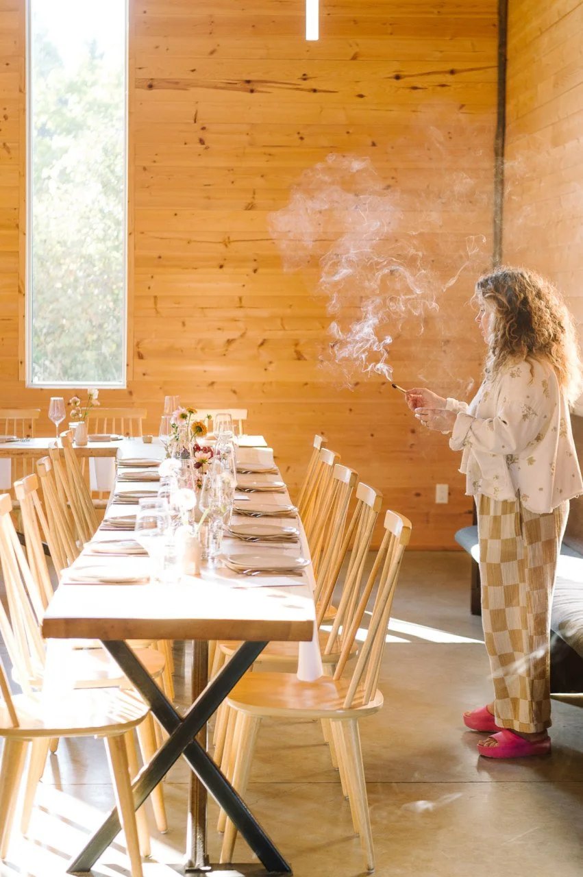 A woman standing in a wooden dining room, holding a lit cigarette with smoke rising, next to a long wooden table set with wine glasses, plates, silverware, and small flower arrangements.