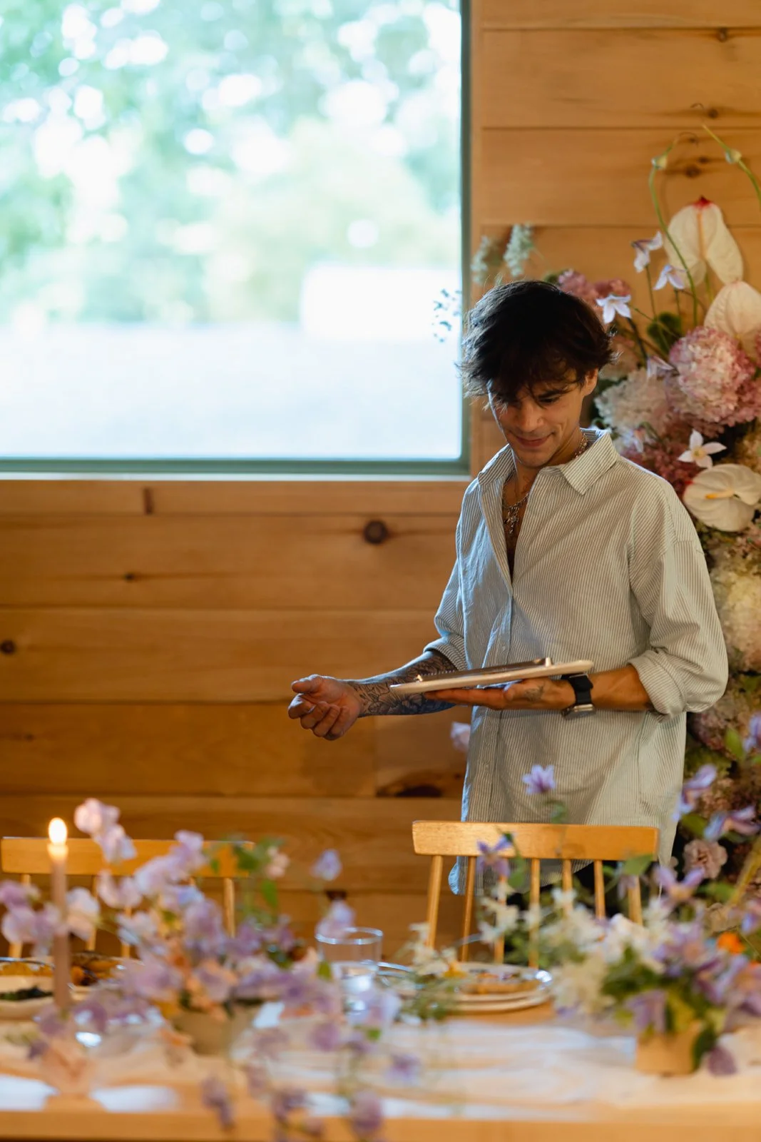 A person with dark hair, tattoos on their arm, wearing a light striped shirt, standing at a table decorated with flowers and candles, inside a wooden-paneled room with a window showing greenery outside.