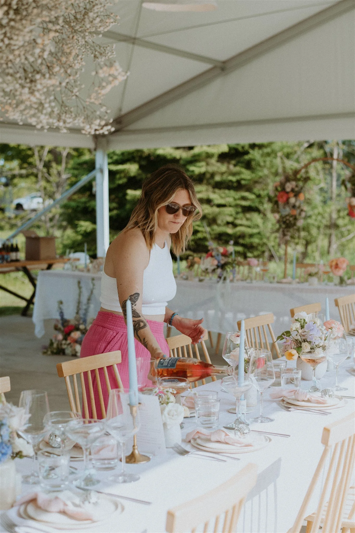 Woman setting up a decorated outdoor event table with flowers, candles, and glassware under a canopy, with trees and flowers in the background.