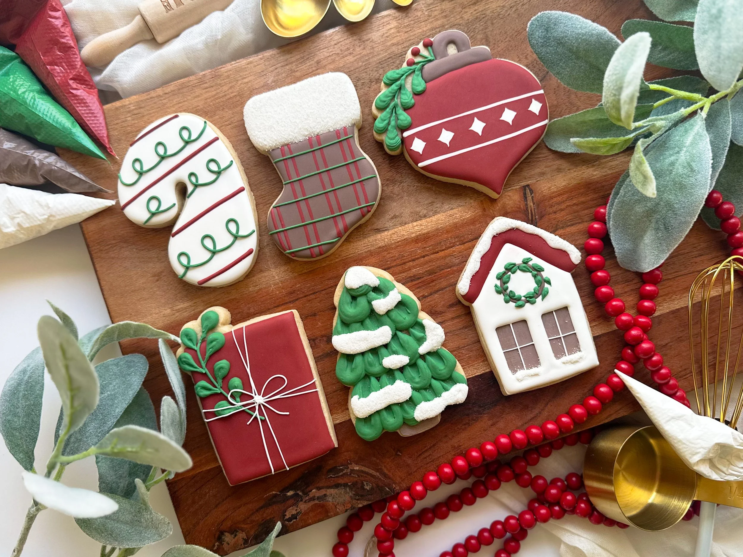 Christmas cookies decorated as candy canes, stockings, ornaments, a house, and a tree on a wooden board surrounded by holiday decorations and greenery.