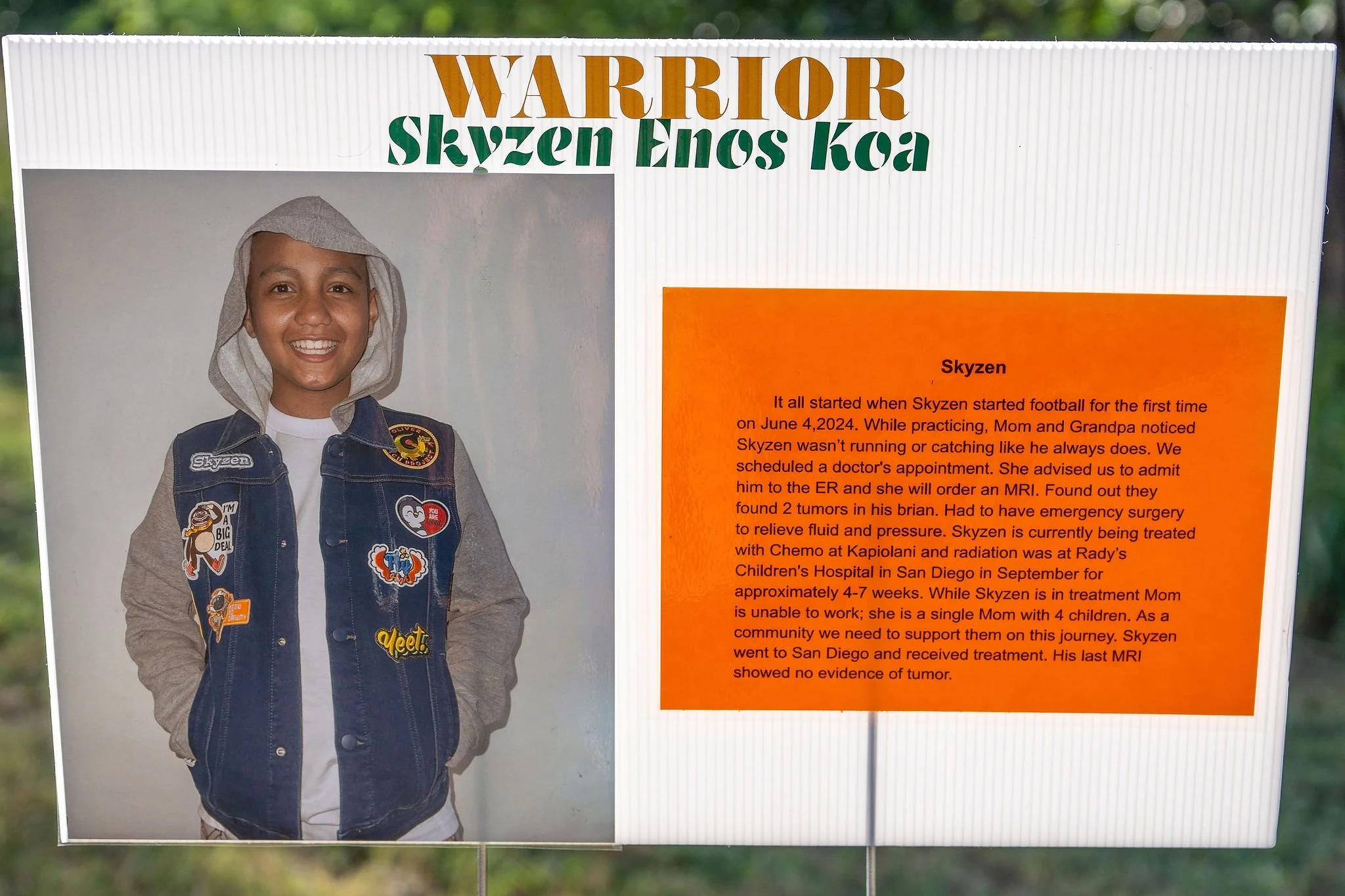 A young boy smiling, wearing a denim vest with various patches and a hoodie. To the right is an orange panel with text about his health journey, mentioning a diagnosis of tumors and treatment at Rady's Children's Hospital.