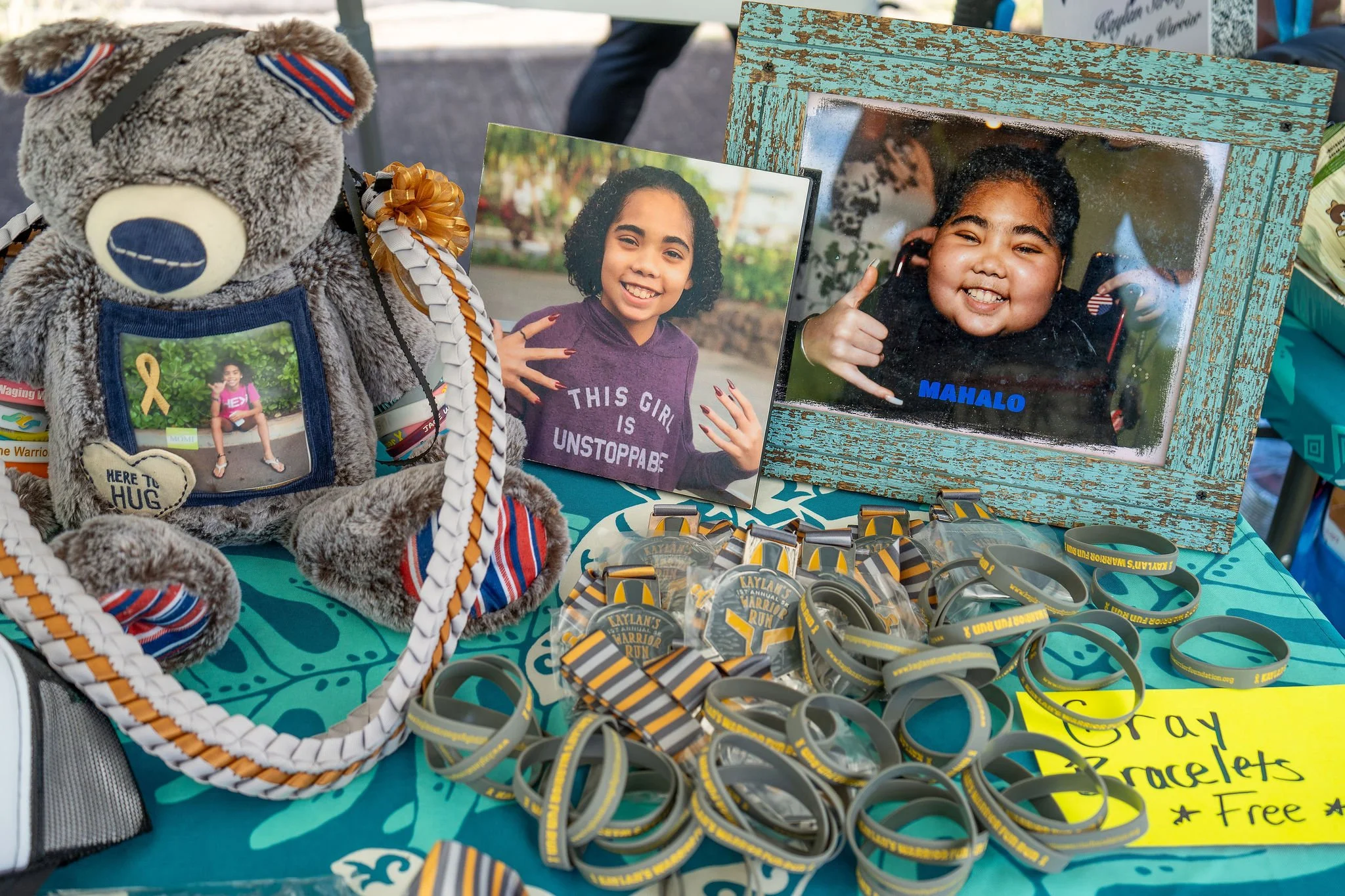 A display table with photos of Kaylan, a young girl, a teddy bear with a photo on its chest, and numerous gray and yellow wristbands. The table has a yellow sign reading 'Gray Bracelets Free.'