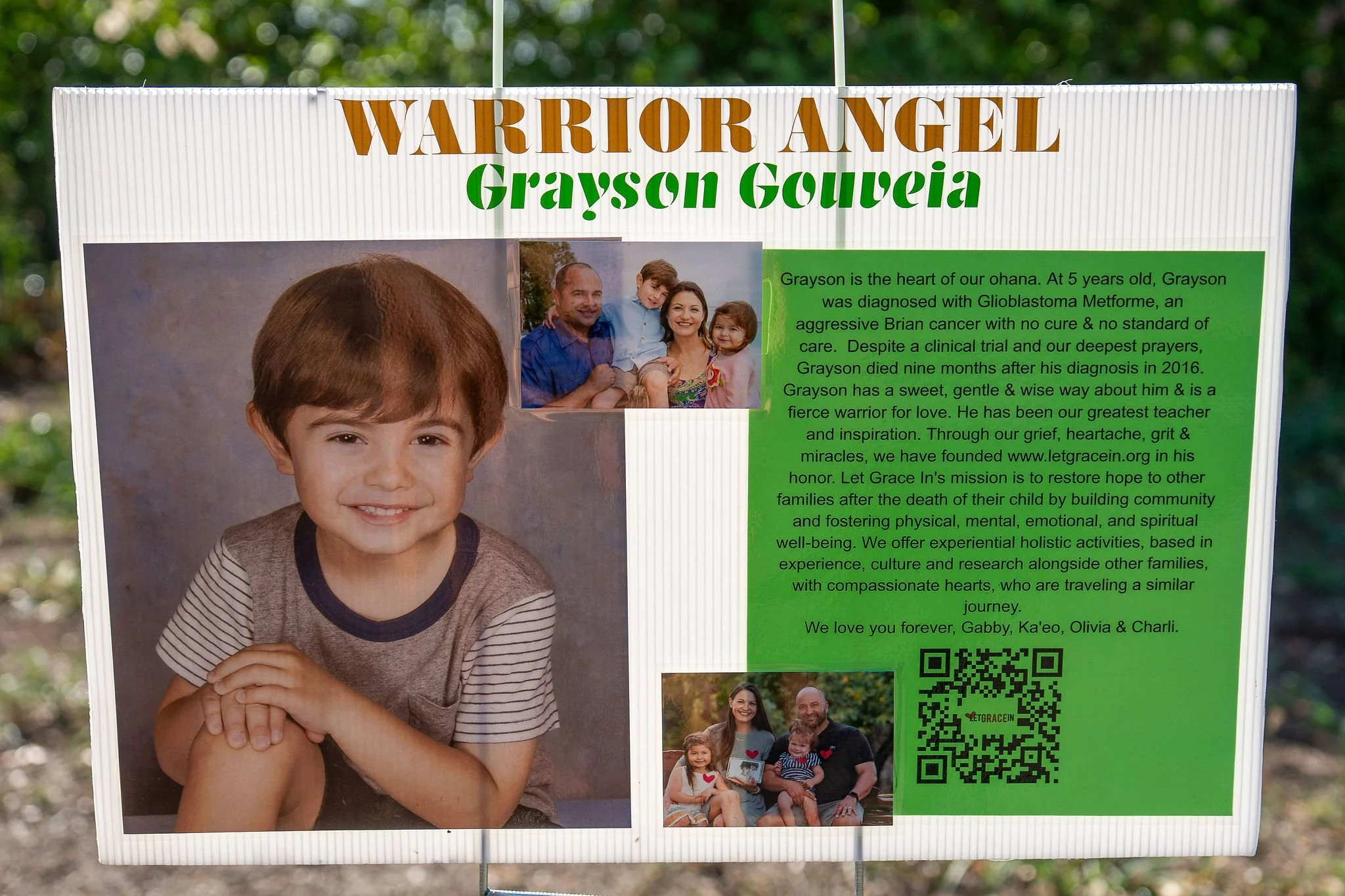 A memorial sign for Grayson Gouveia from Warrior Angel, featuring a large portrait of a young boy with brown hair and a smile, along with smaller photos of a family and a couple with children. The sign includes a detailed tribute about Grayson, menti