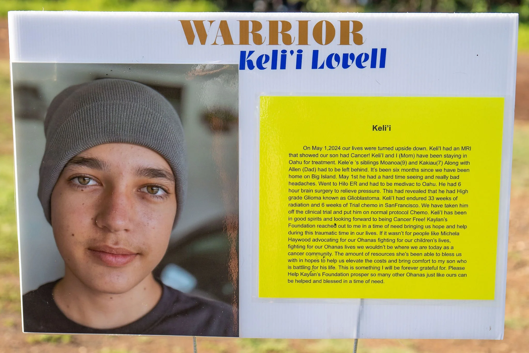 A memorial display with a large photograph of a young boy wearing a gray beanie, and a yellow poster explaining her health struggles and treatment history, with the title 'WARRIOR' and his name 'Keli'i Lovell.'