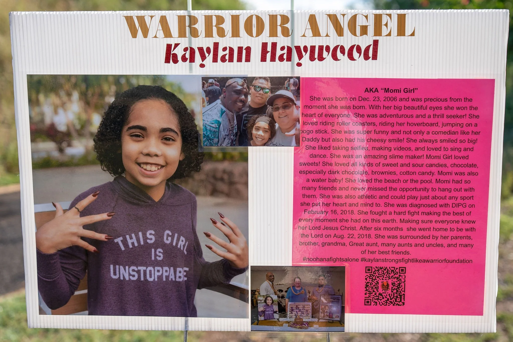 A memorial display for Kaylan Haywood, AKA 'Momi Girl,' featuring a large photo of a smiling young girl with curly hair, wearing a purple shirt that reads 'This Girl Is Unstoppable.' The display includes smaller pictures of her with family, a pink bi