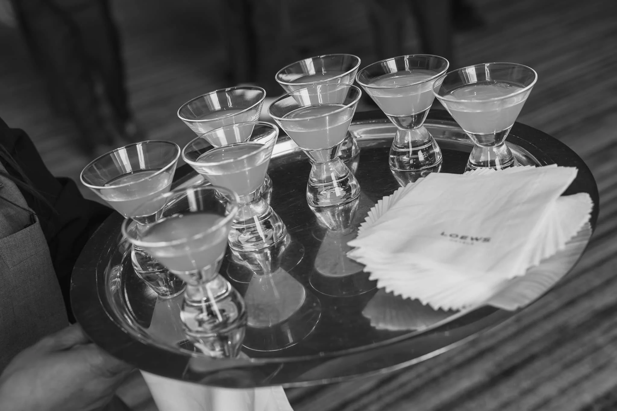Black-and-white photo of a server holding a tray of cocktails in stemmed glasses with napkins at a wedding