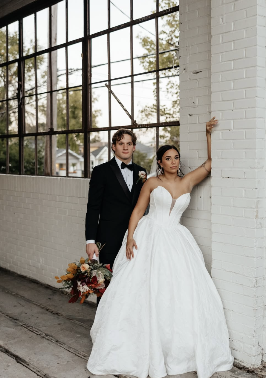 Modern fine-art wedding photo of a couple in elegant attire against a white-brick backdrop, combining editorial style with authentic emotion.