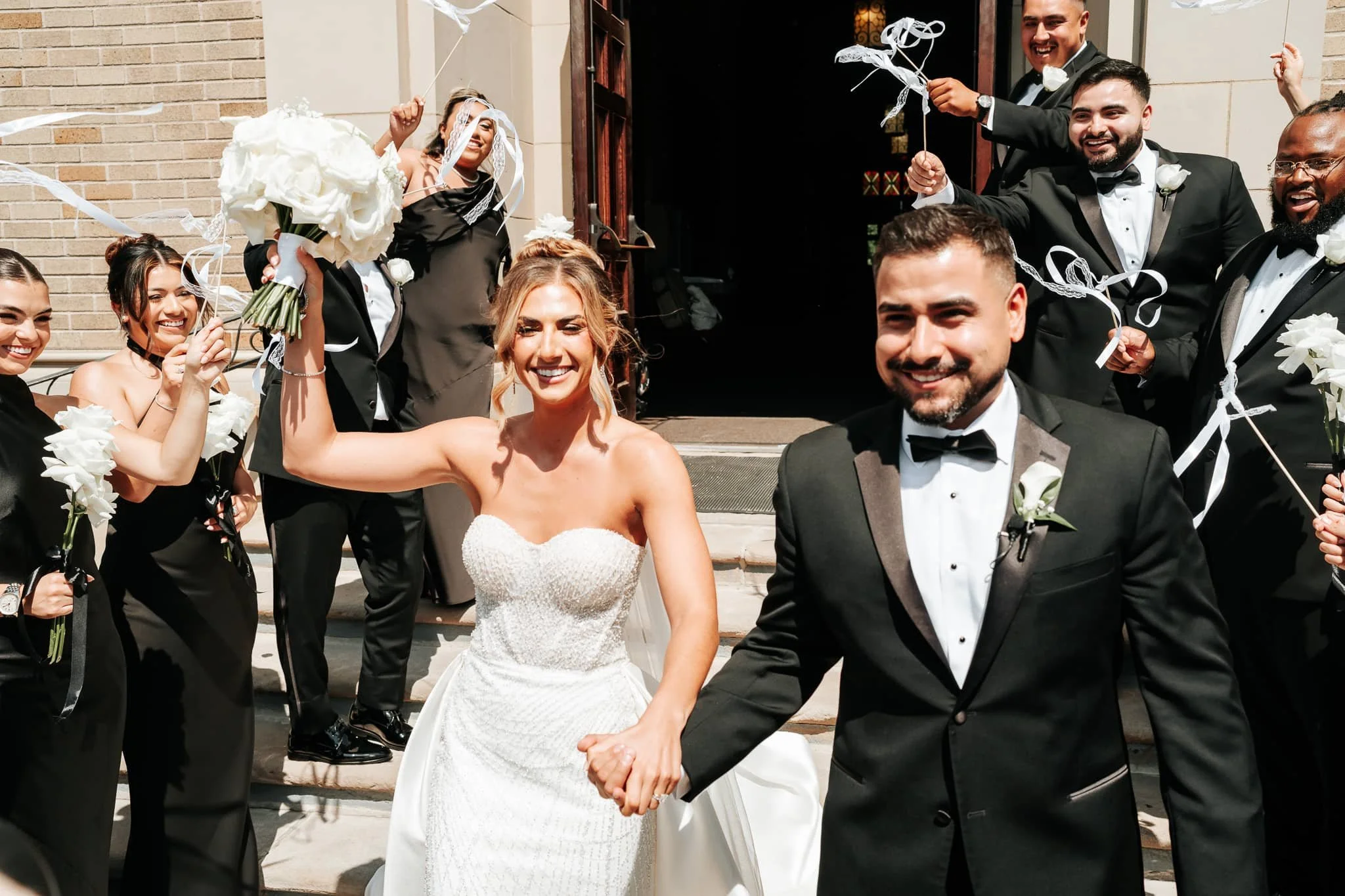Bride and groom walking hand in hand down church steps while their wedding party cheers and waves ribbon wands