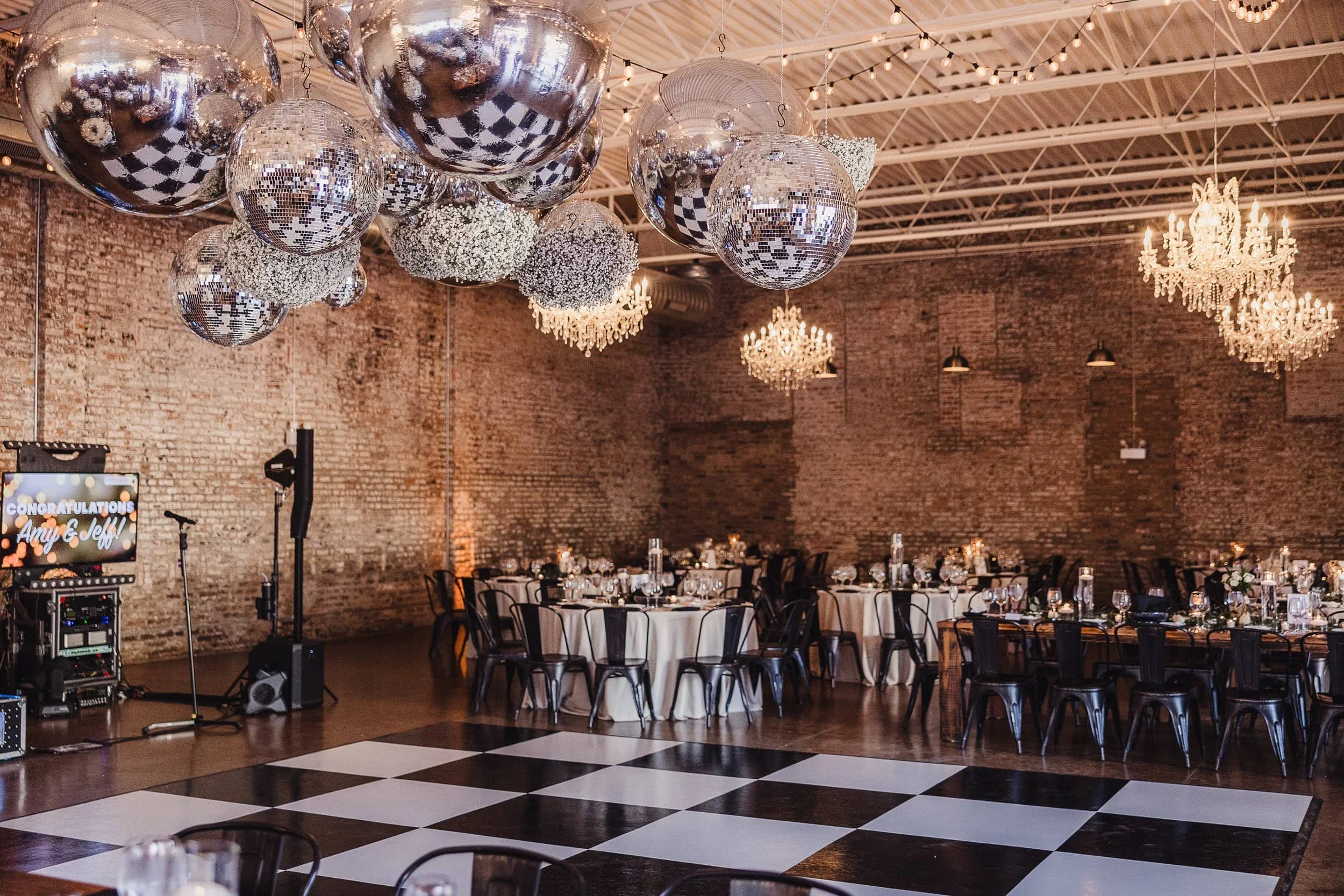 A decorated event space with multiple large disco balls and chandeliers hanging from the ceiling, round tables with white tablecloths and black chairs, brick walls, and a checkered dance floor also visible.