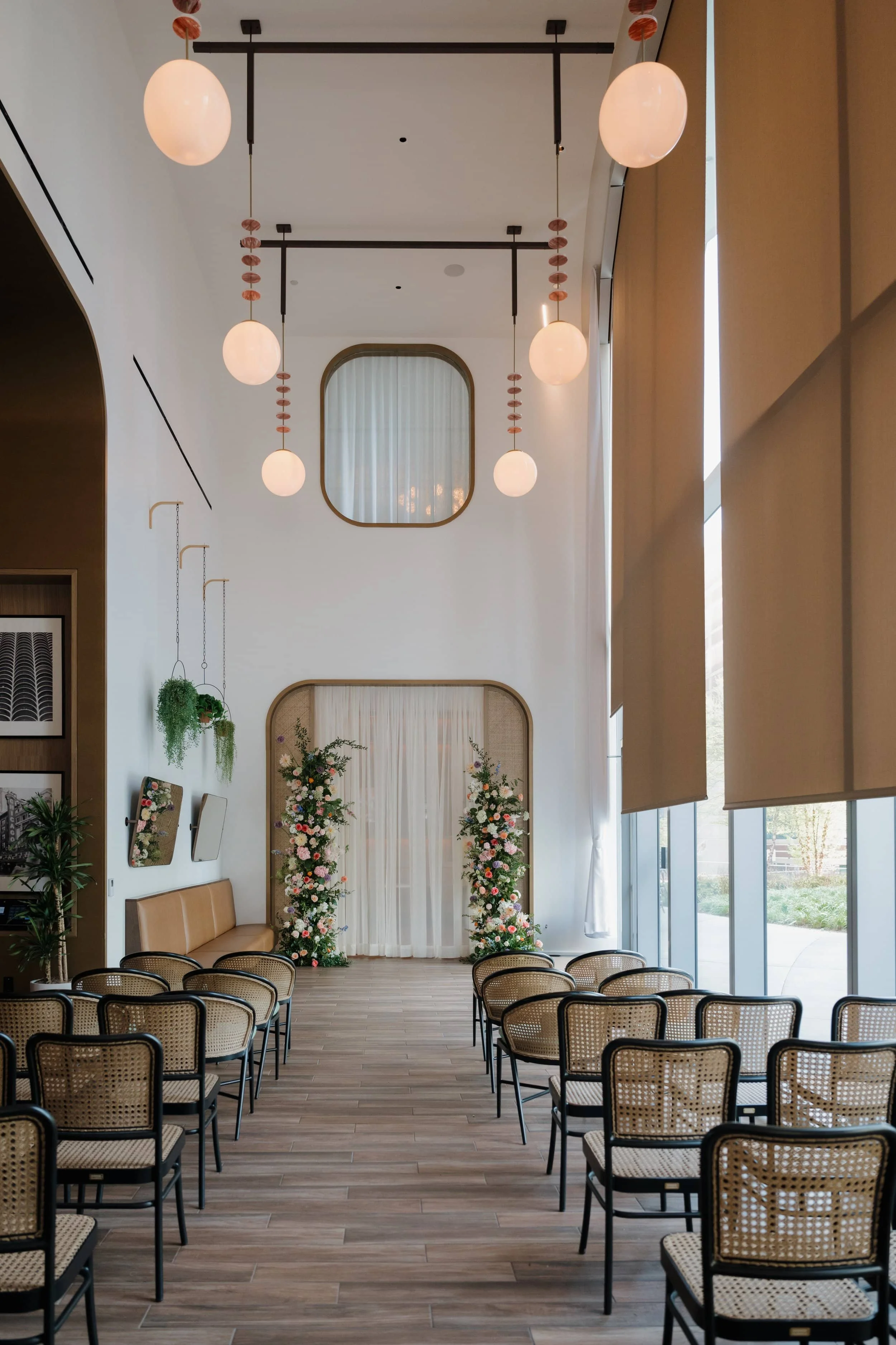 An indoor space set up for a ceremony with rows of chairs facing a decorated backdrop with floral arrangements and a white curtain. The room has large windows with beige blinds, modern lighting fixtures hanging from the ceiling, and some wall decorations including framed art and hanging plants.