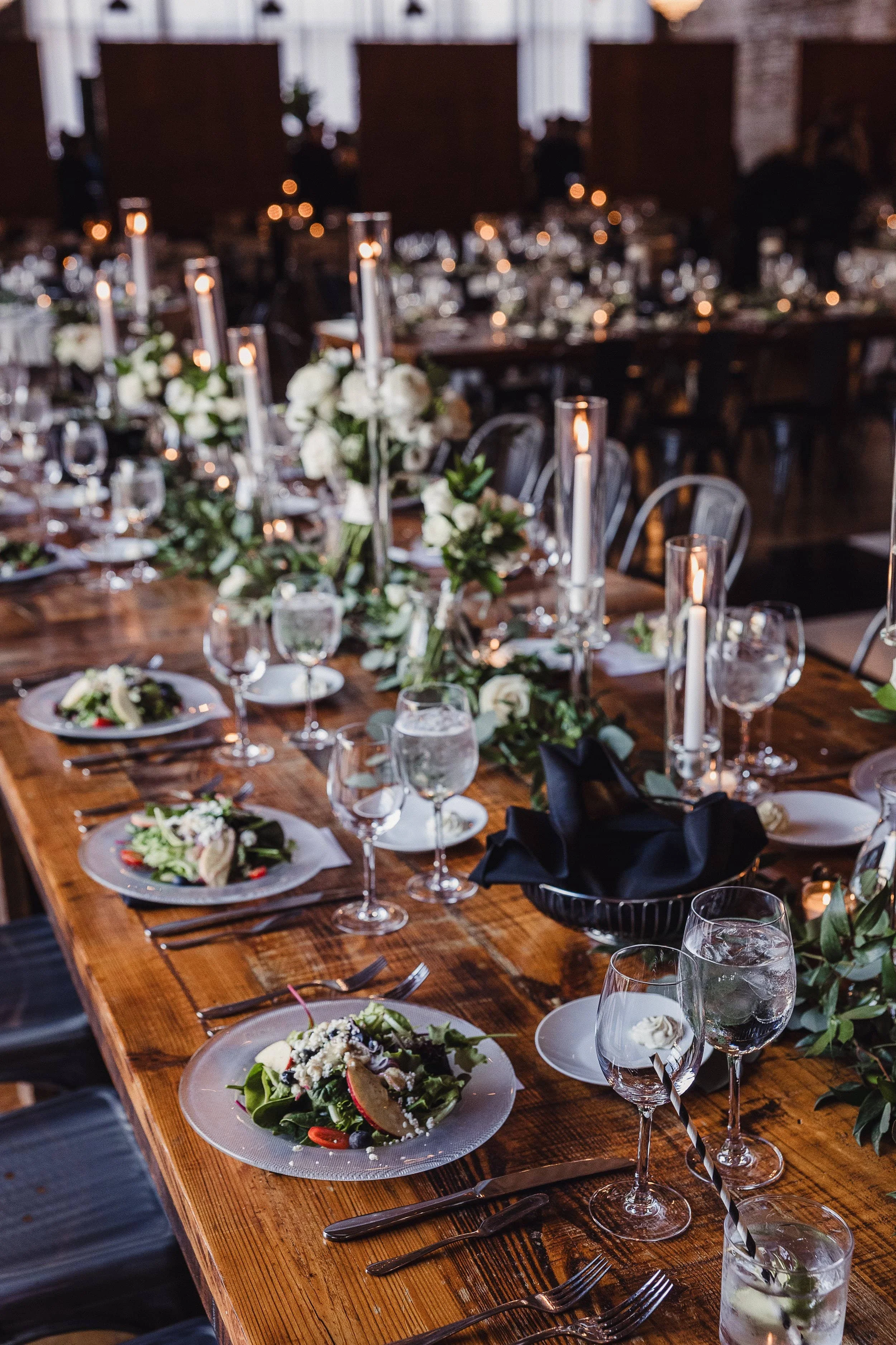 A long wooden dining table set for a meal with salad plates, wine glasses, and black napkins. The table is decorated with white flower arrangements and tall, lit candles. In the background, there are more tables and chairs, creating an elegant, cozy ambiance.