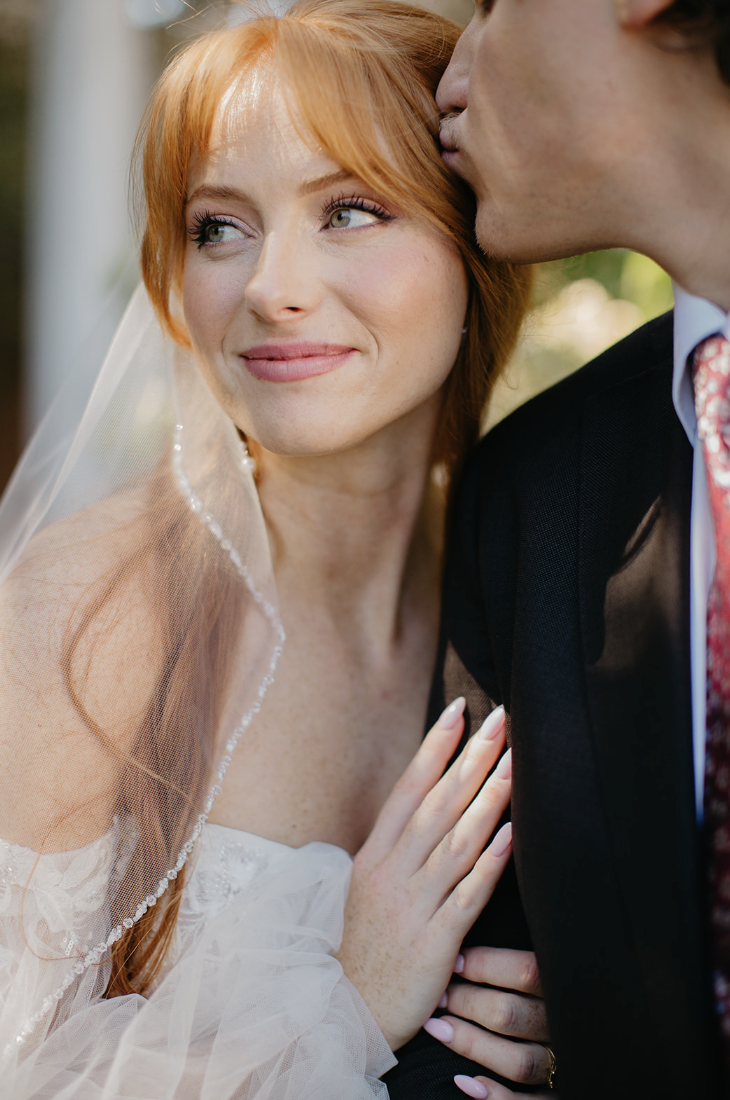 Bride smiling while groom kisses her forehead, emotional fine-art wedding photo in natural light.