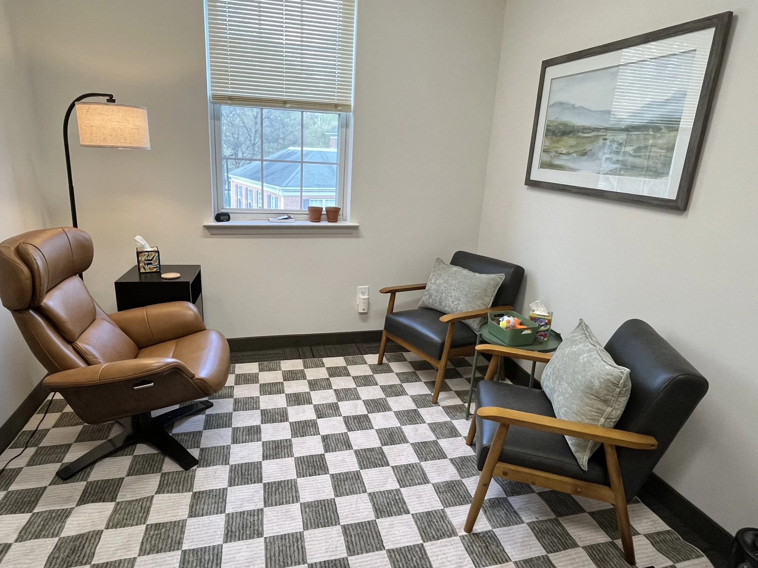 Office space Hopeful Hearts Therapy in Princeton, NJ. There is a seating area for therapy. There are two matching grey chairs, a brown chair, a lamp with warm lighting, and a picture on the wall of a peaceful landscape. 