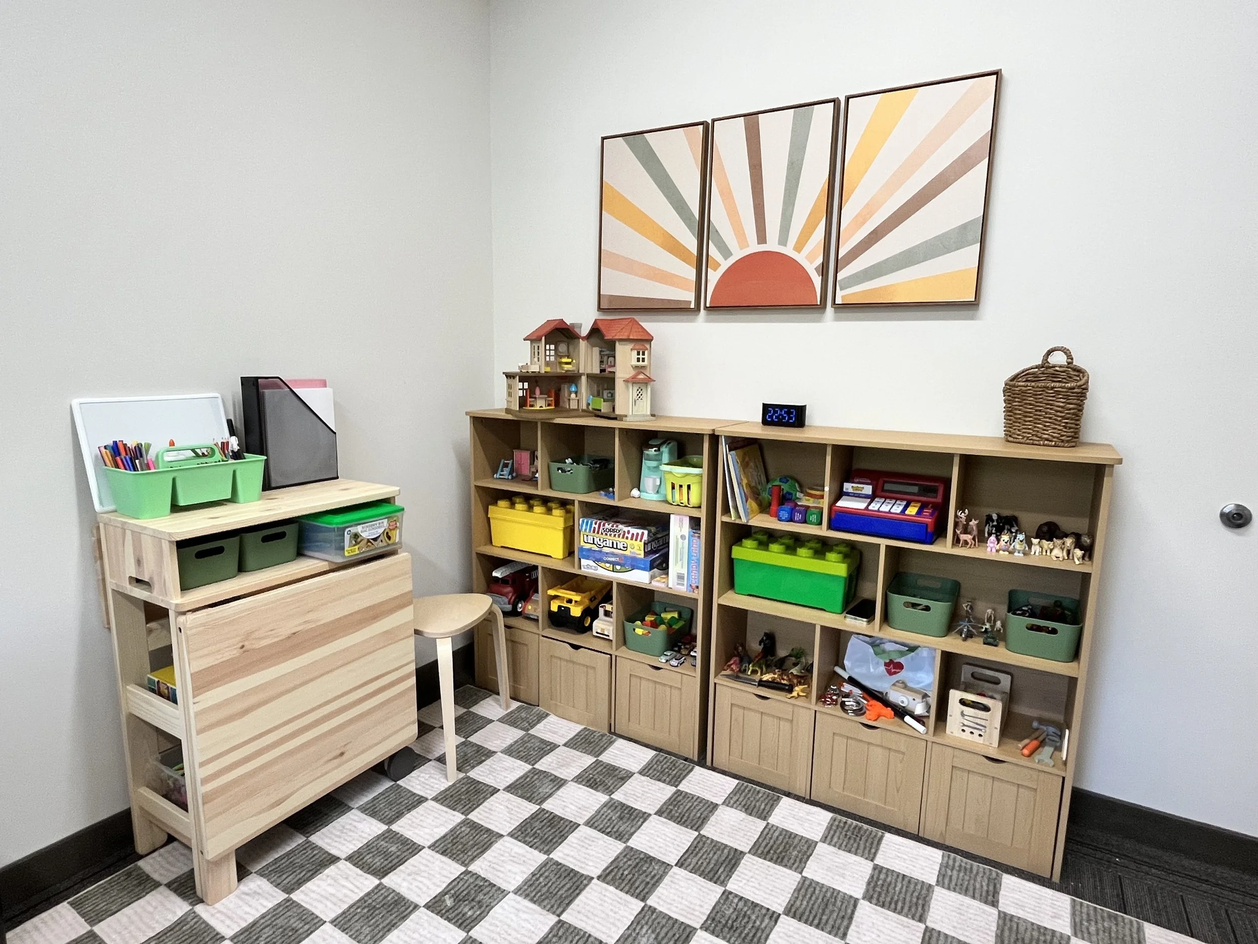 Office space Hopeful Hearts Therapy in Princeton, NJ.  A children's play area in an office space. There are shelves with various toys, a wooden cart with art supplies, and a picture of a colorful sun on the wall above.