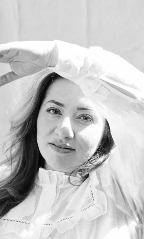 Black and white photo of a woman with long hair, wearing a light-colored blouse, smiling, with her hand resting on her forehead.