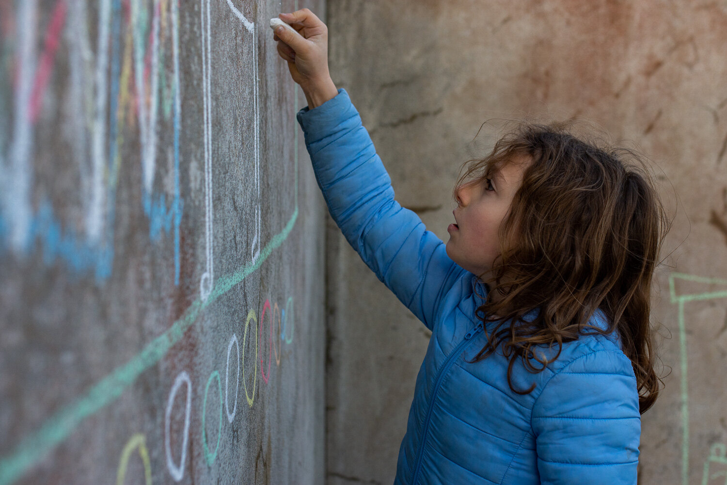 Girl with curls blue coat drawing on wall with chalk