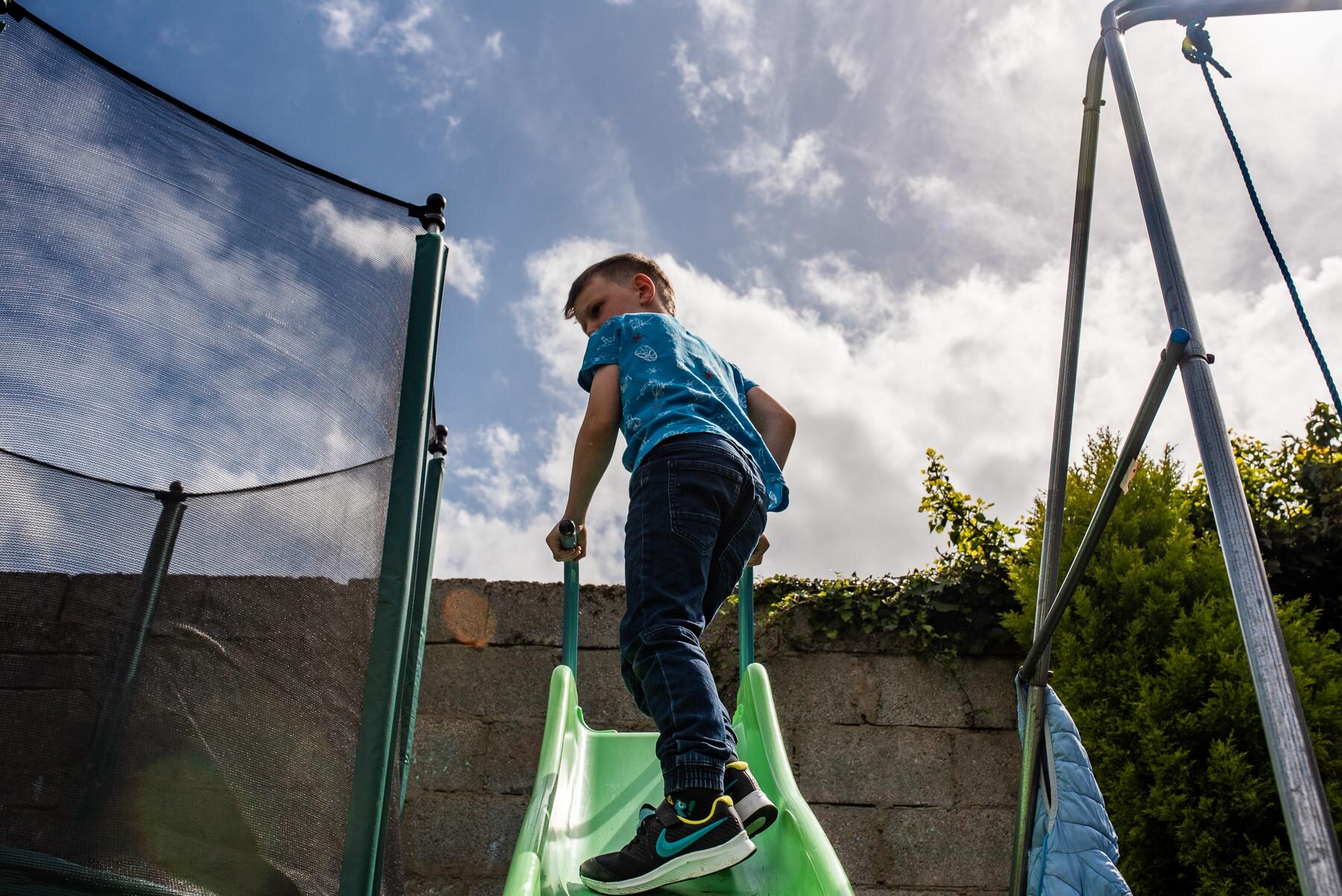 boy standing on green slide next to trampoline and swing
