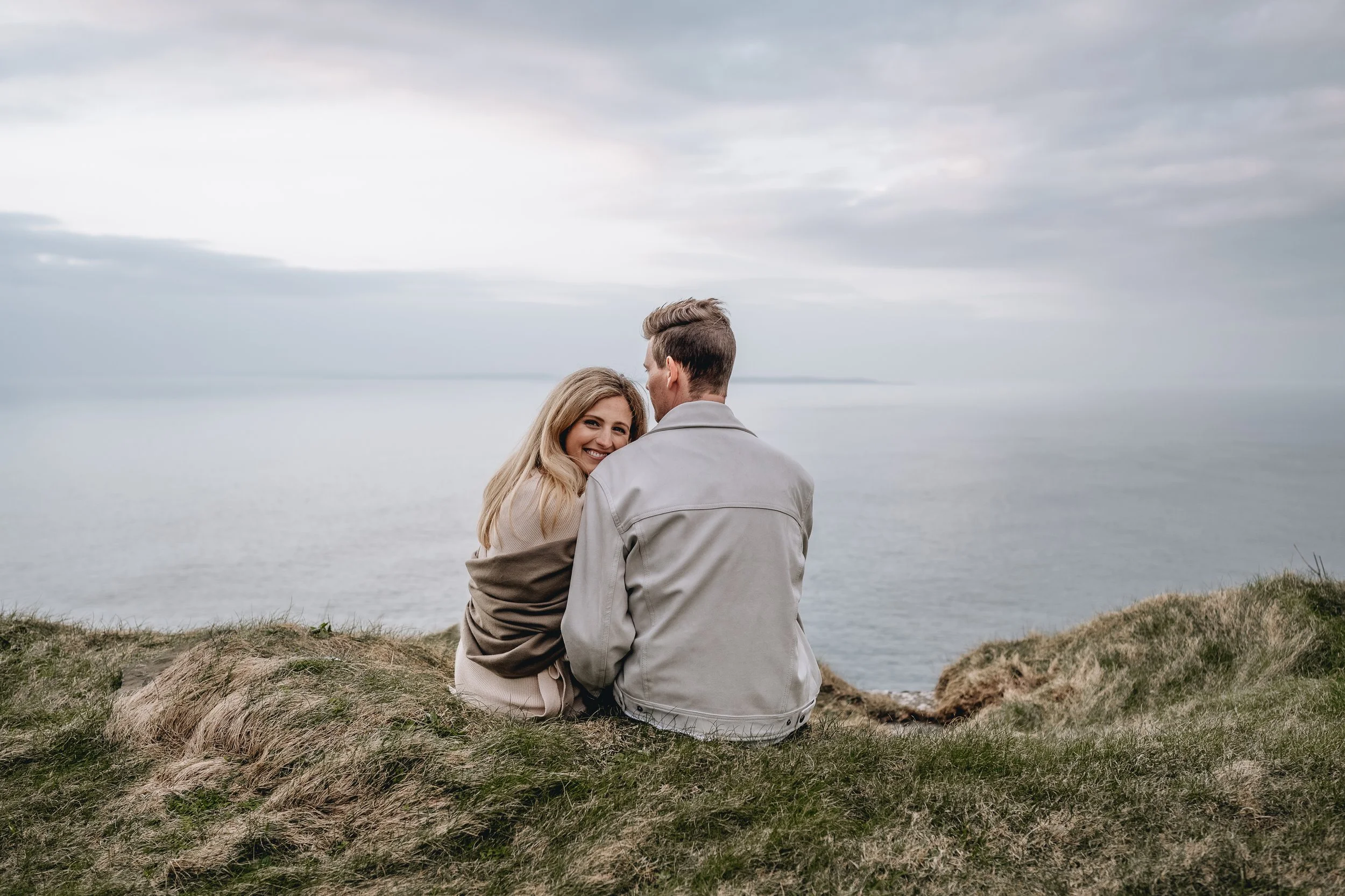 couple sitting on cliff edge blond woman looking over man's shoulder at the Cliffs of Moher photo session photographer ireland