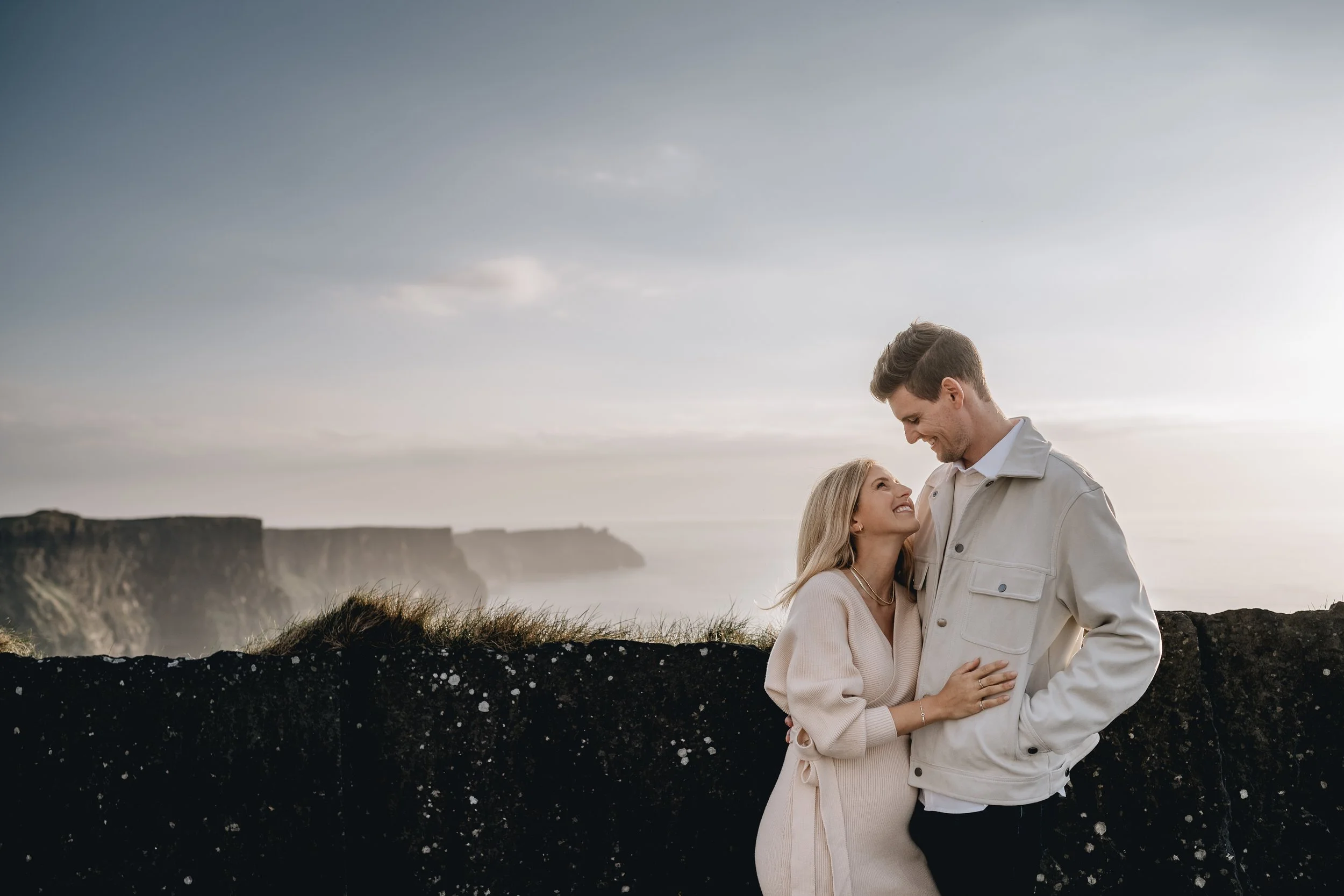 pregnant woman smiling at husband standing in front of the Cliffs of Moher during maternity photos Ireland