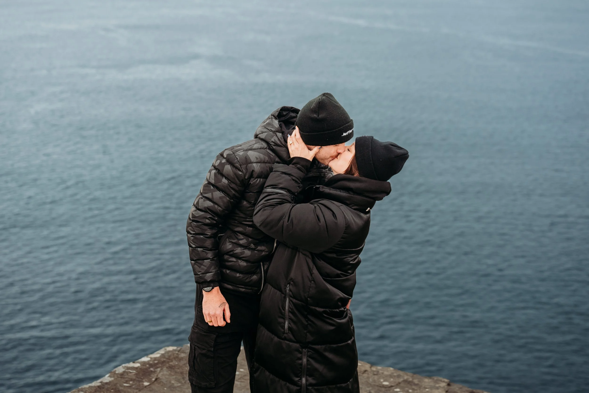 Couple dressed in black kisses with ocean background cliffs of mother session photographer ireland Marie I'Mahony