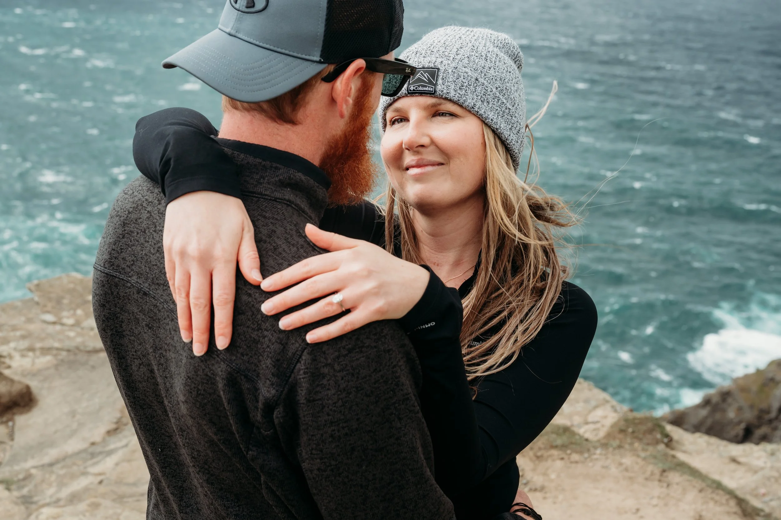 woman gazes at man in hat on top of cliff ocean behind them couples photo session photographer Marie O'Mahony