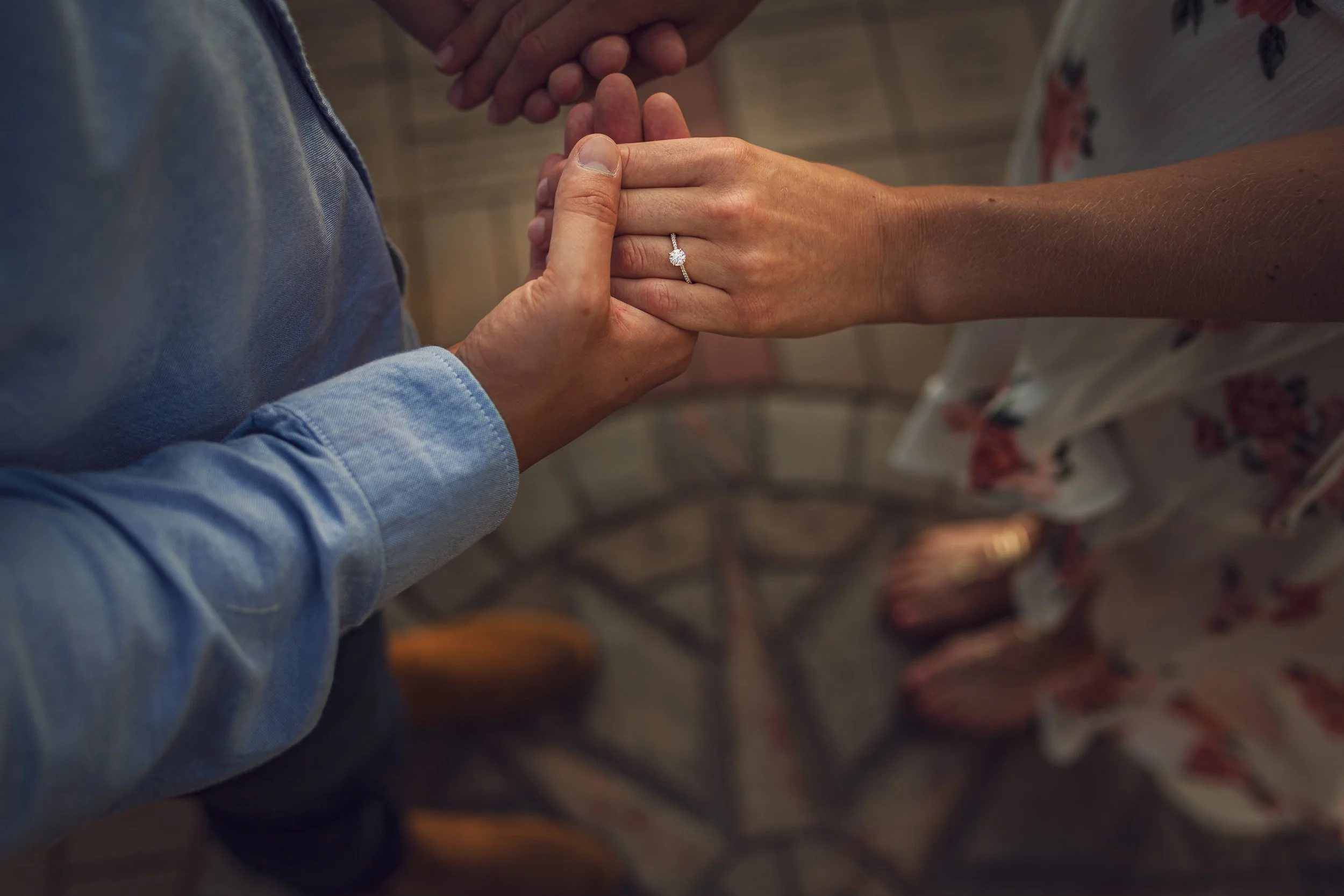 Close-up of a couple holding hands, with the woman wearing a diamond engagement ring, indoors.