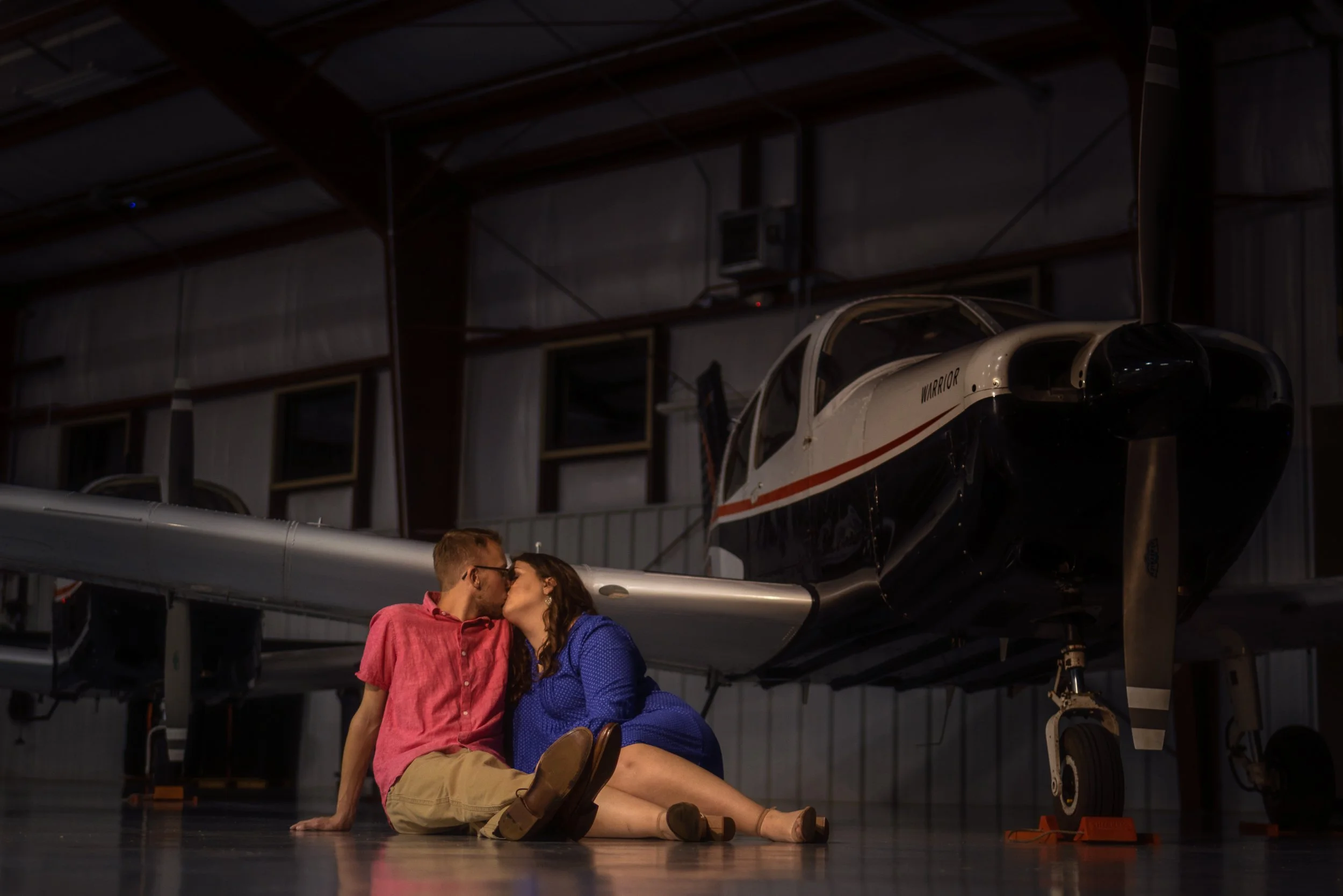 A couple sitting on the floor inside a hangar, sharing a kiss in front of a small aircraft, with a man in a red shirt and woman in a blue dress.