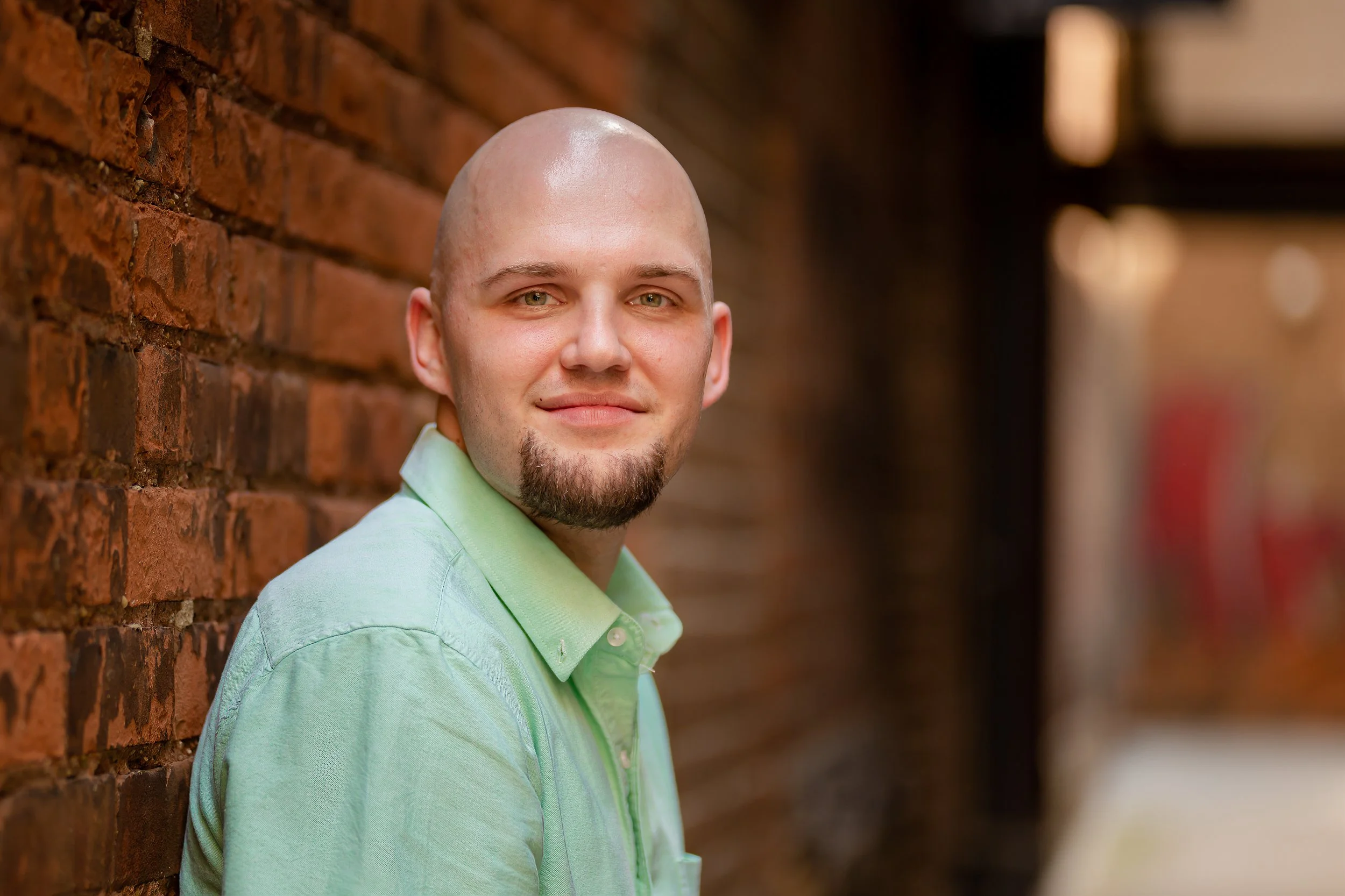 A man with a shaved head and green eyes, wearing a light green shirt, standing outdoors against a brick wall, smiling at the camera.
