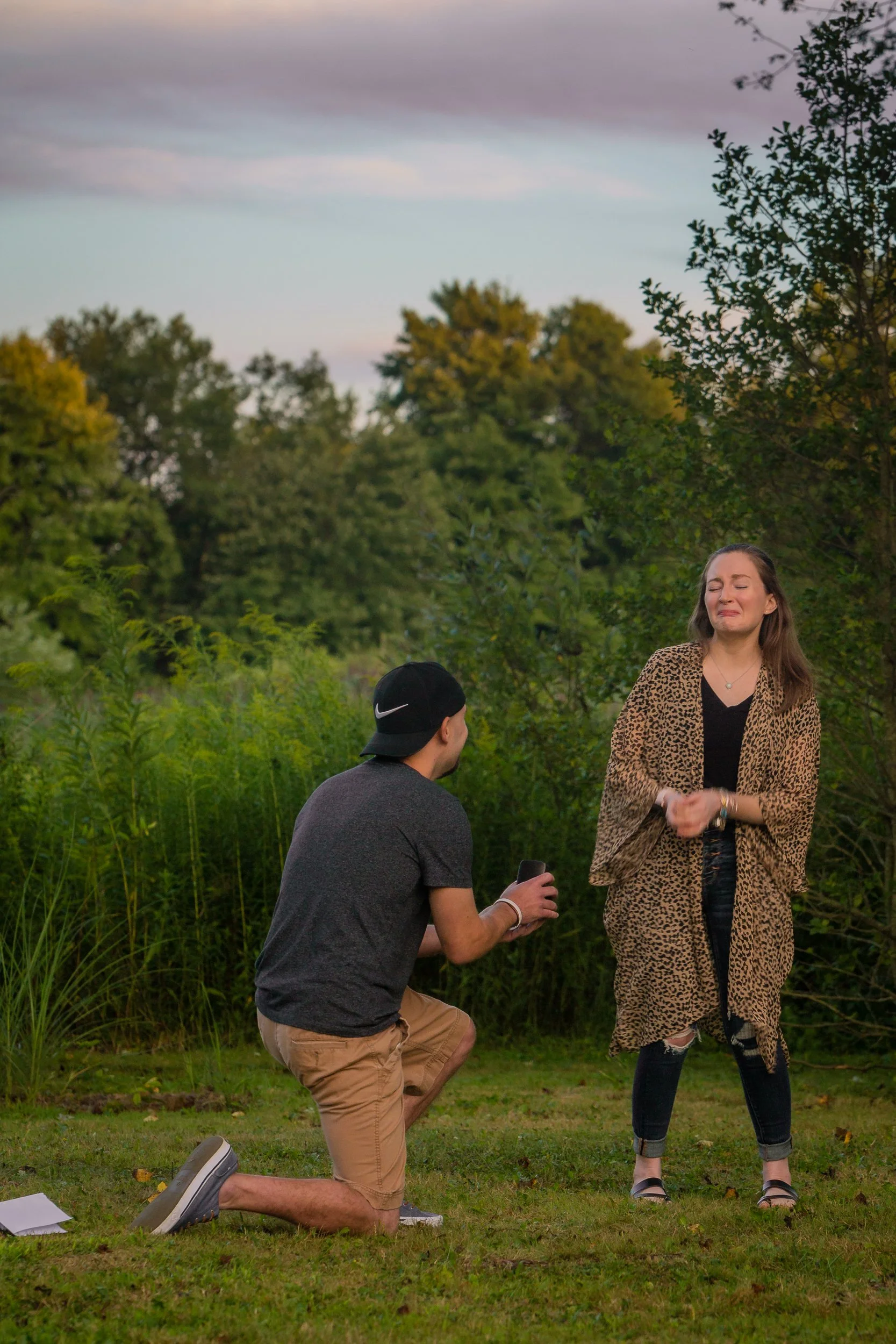 A man on one knee proposing to a woman in an outdoor setting with greenery and trees, she appears emotional.