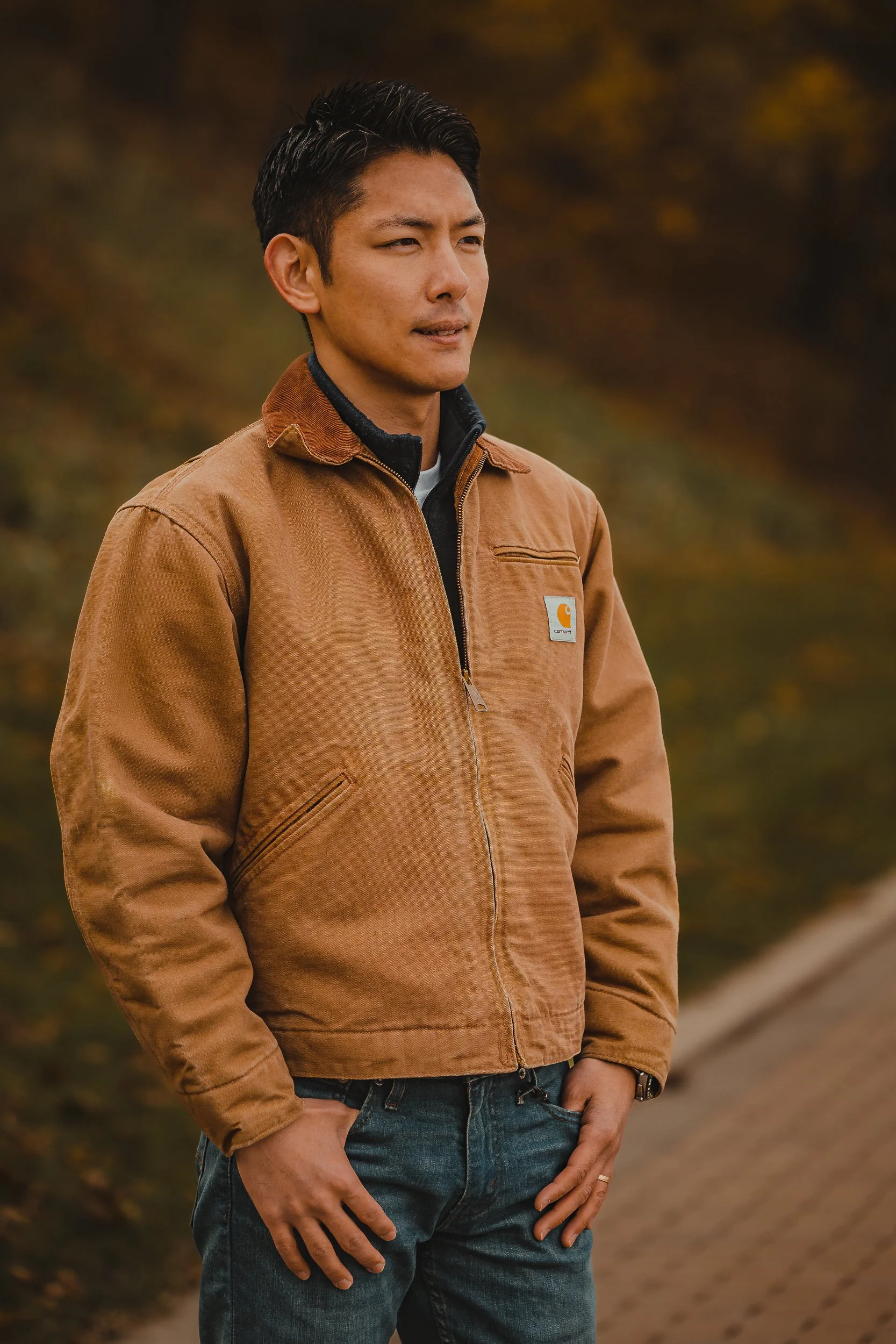 A man standing outdoors, wearing a brown Carhartt jacket, with one hand resting on his pocket and the other hanging by his side, with a blurred background of autumn foliage.