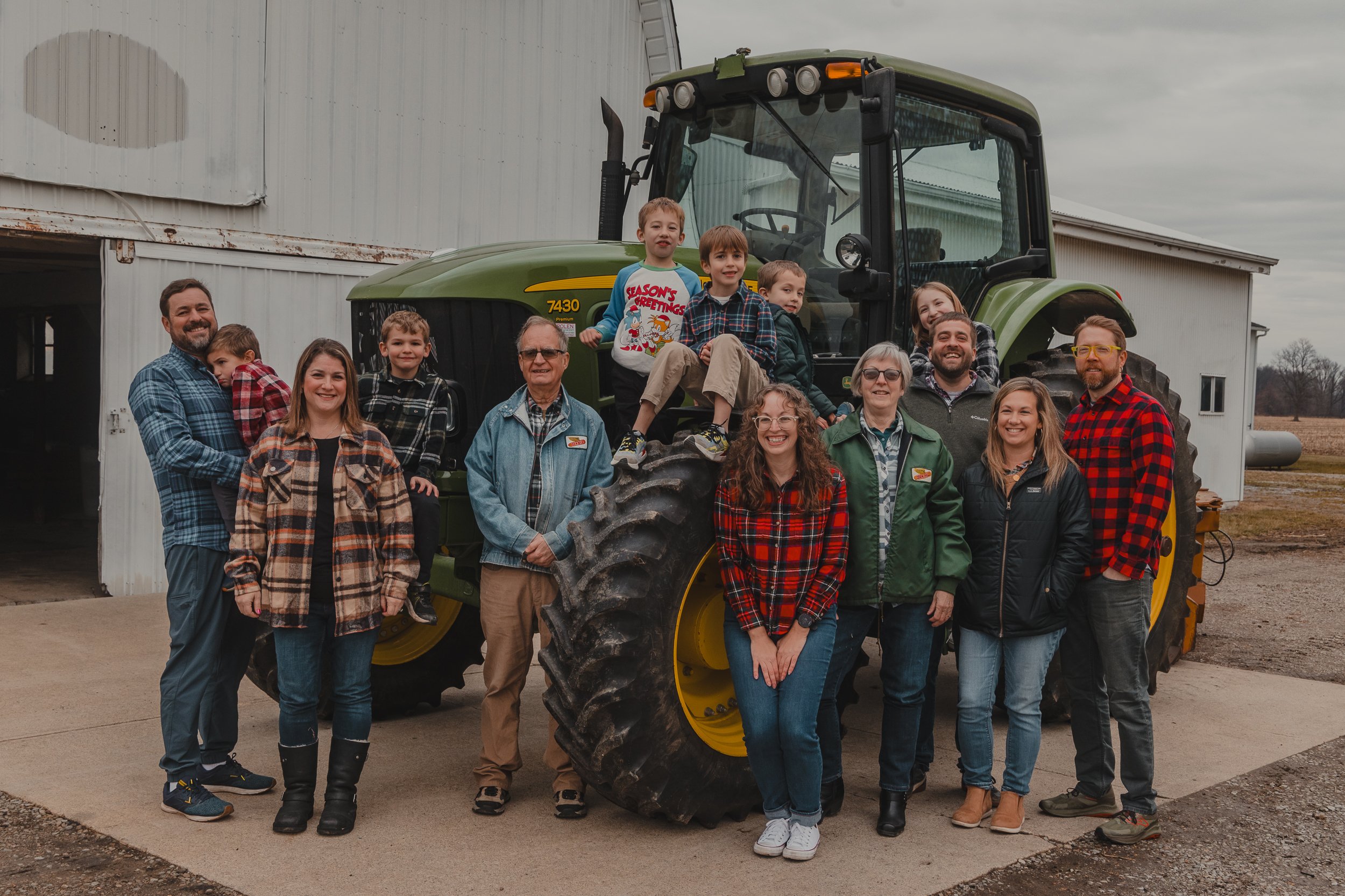 A group of 13 people, consisting of children, teenagers, and adults, gathered around a large green farm tractor outside a white barn with a cloudy sky in the background.