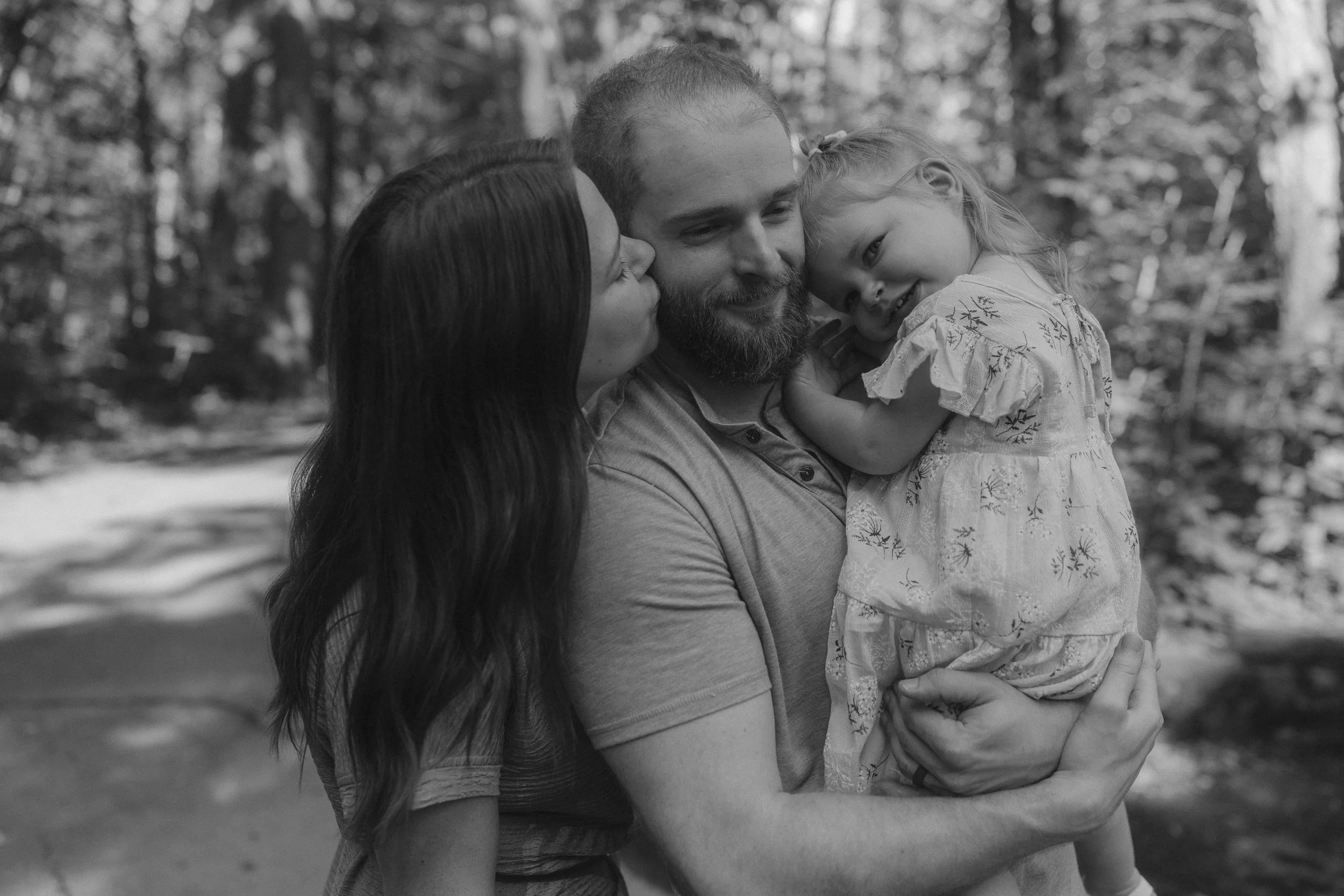 A black and white photo of a man holding and cuddling two young girls, one girl is kissing the man's cheek, and the other girl is resting her head on his shoulder. They are outdoors surrounded by trees.