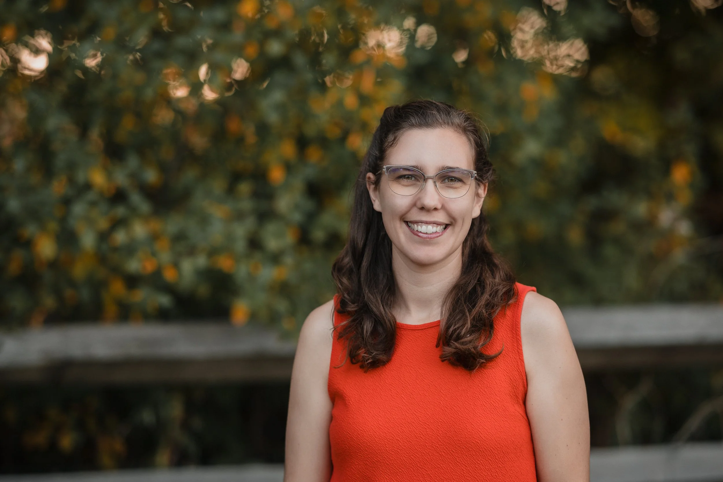 A smiling young woman with brown, curly hair and glasses, wearing an orange sleeveless top, standing outdoors with blurred green and yellow foliage in the background.