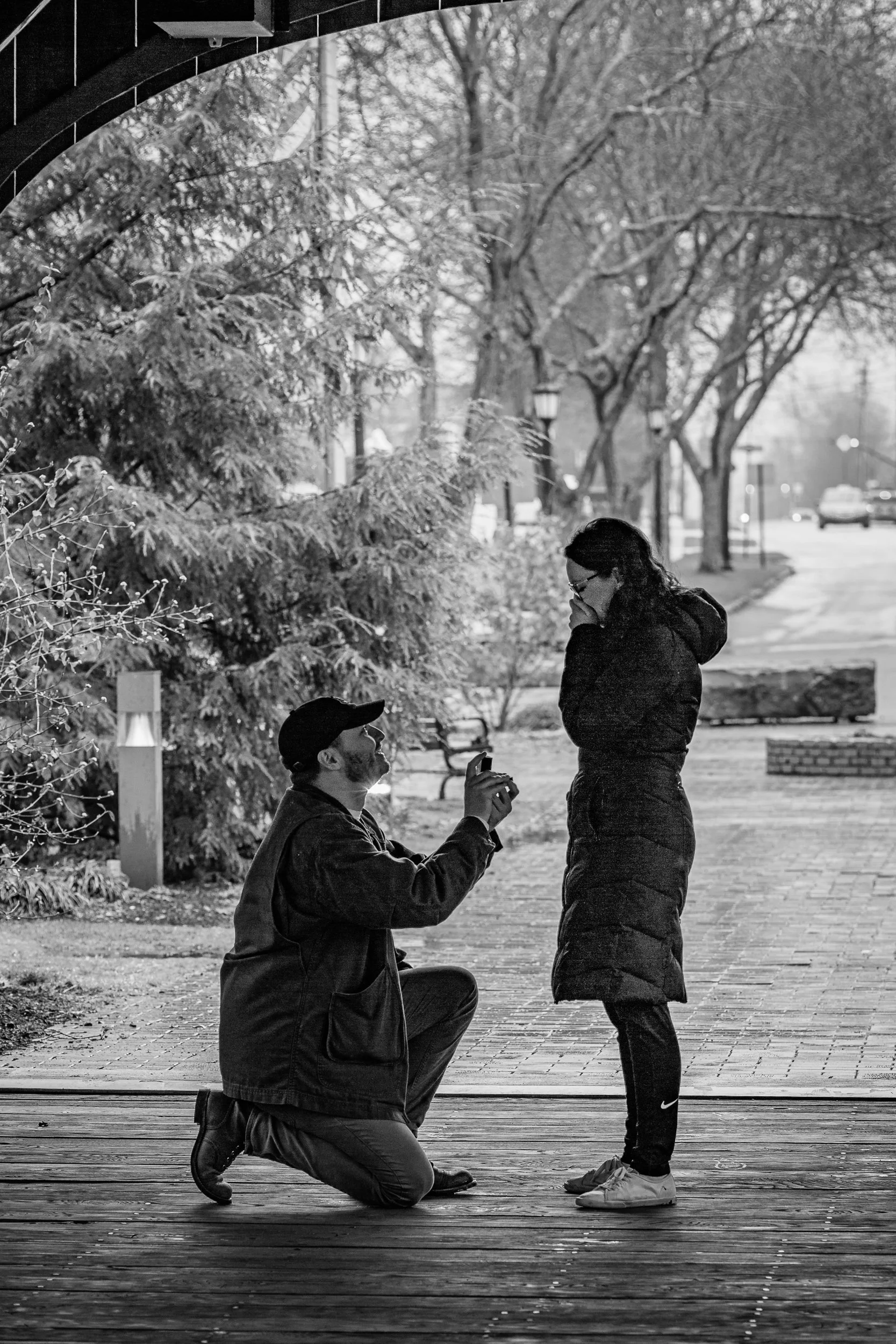 A man kneeling on a wooden deck proposing to a woman, who appears emotional and is covering her mouth with her hand, outdoors in a park-like setting with trees and lamp posts.