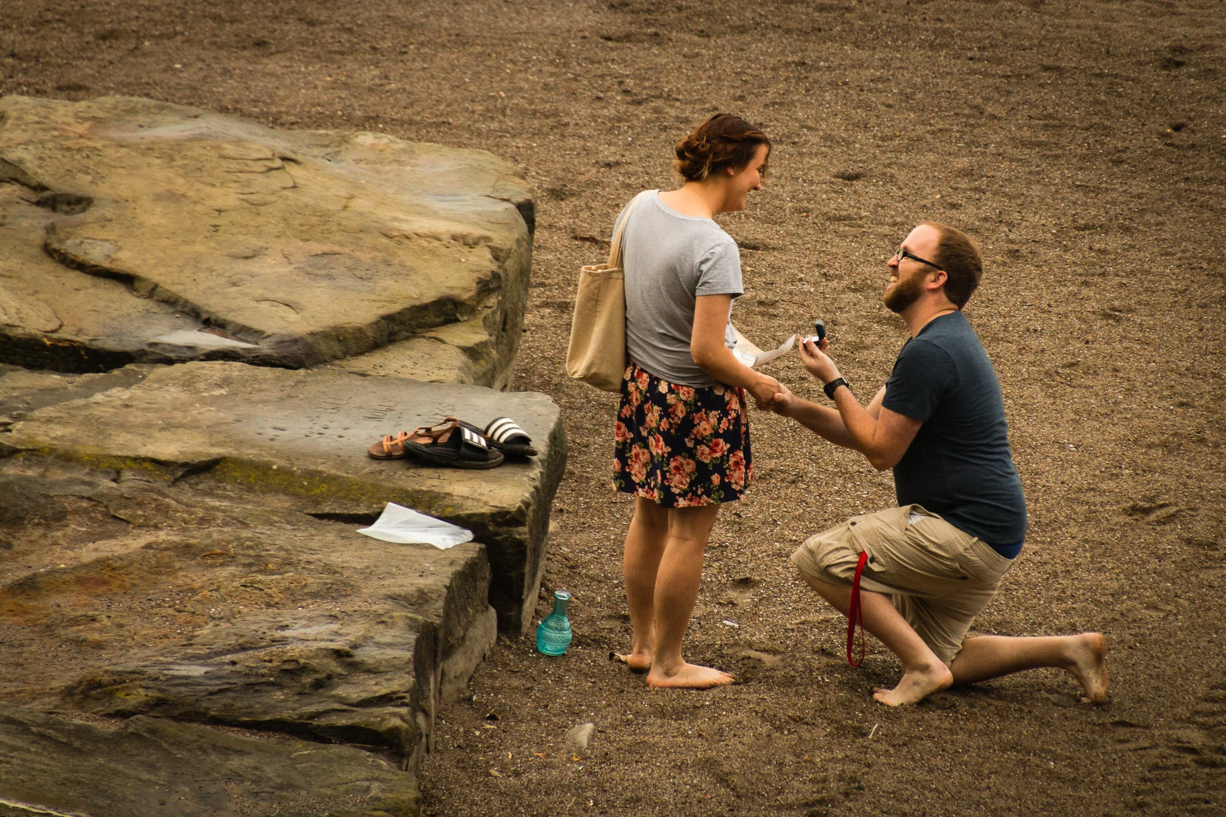 A man on one knee proposing marriage to a woman on a beach, with a large rock formation nearby.