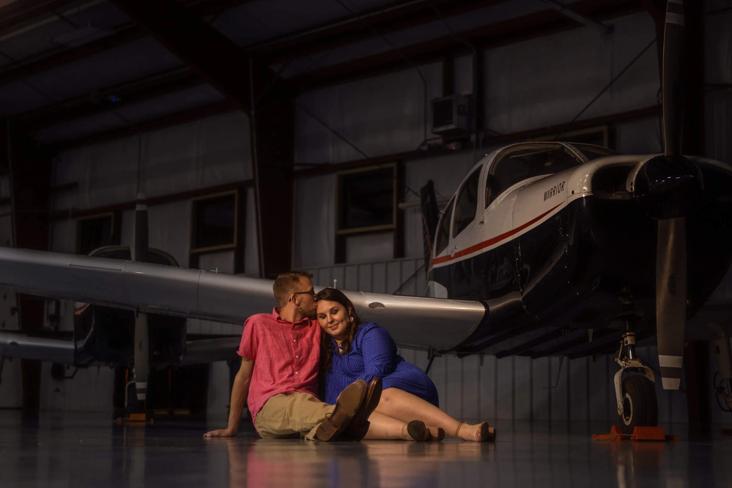 engagement session, couple sitting on ground in front of trainer airplane, pink shirt, blue shirt, dressed nicely, bride to be