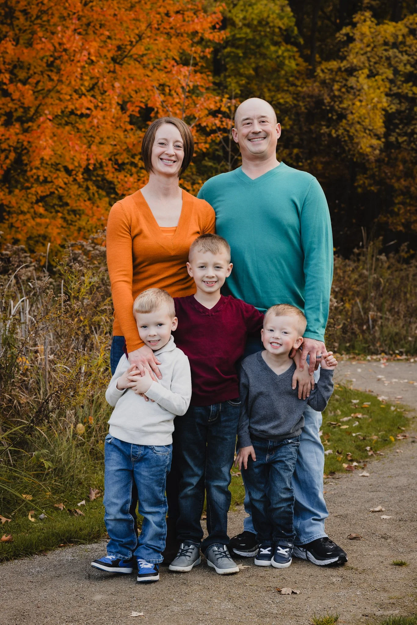 A family of five standing outdoors during autumn, with colorful fall foliage in the background. The family includes a woman, man, and three young boys, all smiling at the camera.