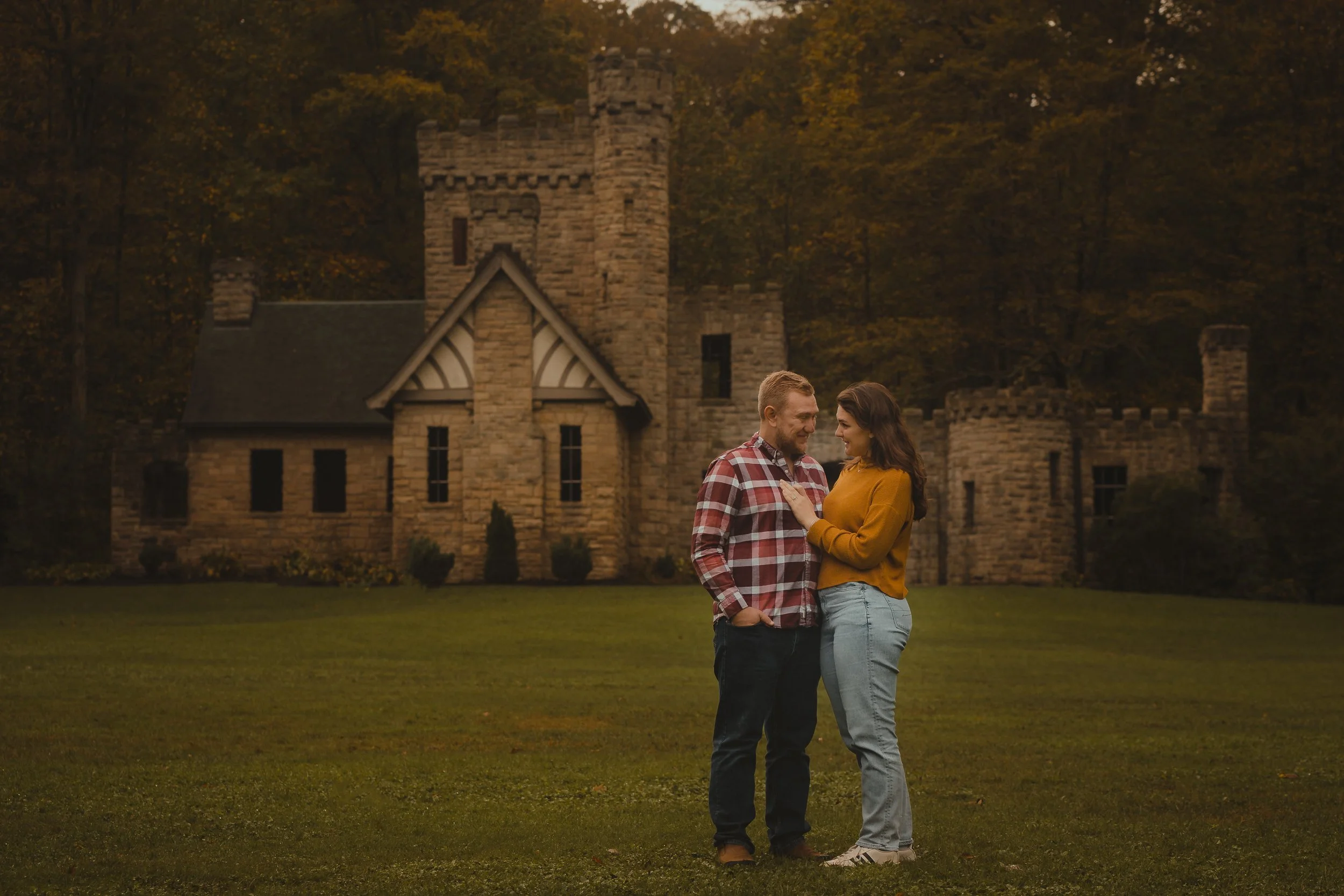 A man and woman standing close together on a lawn in front of a castle-shaped building during dusk, smiling and looking at each other.