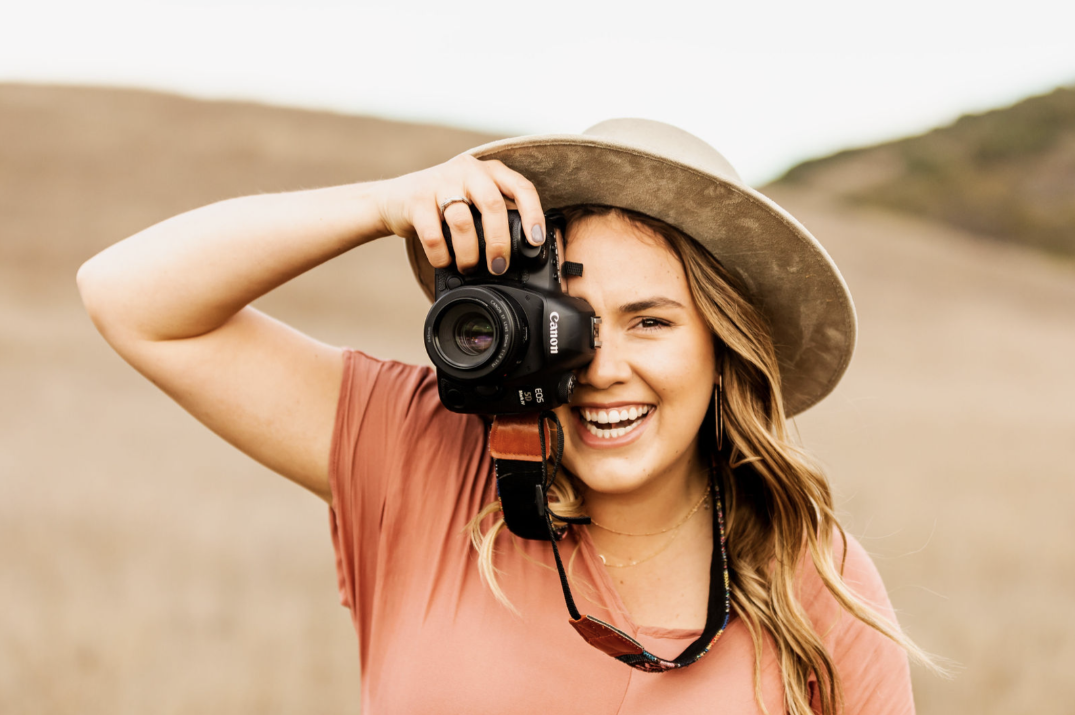 A woman holding a Canon camera up to her face while outdoors, wearing a wide-brimmed hat and a reddish-brown shirt.