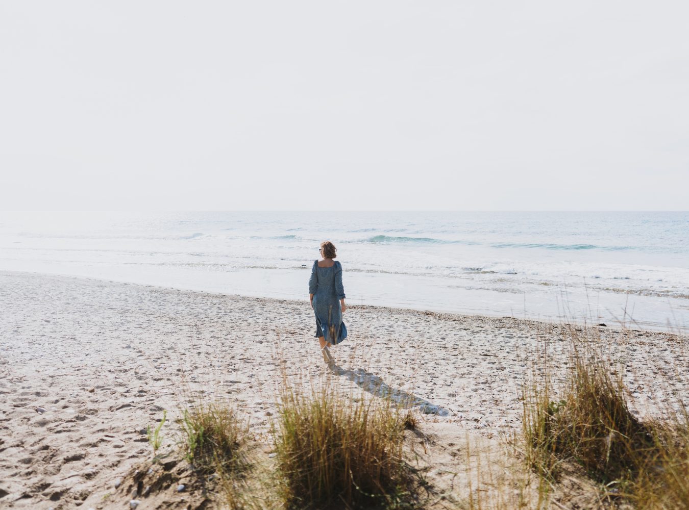 En person i en blå klänning går på en sandstrand mot havet.