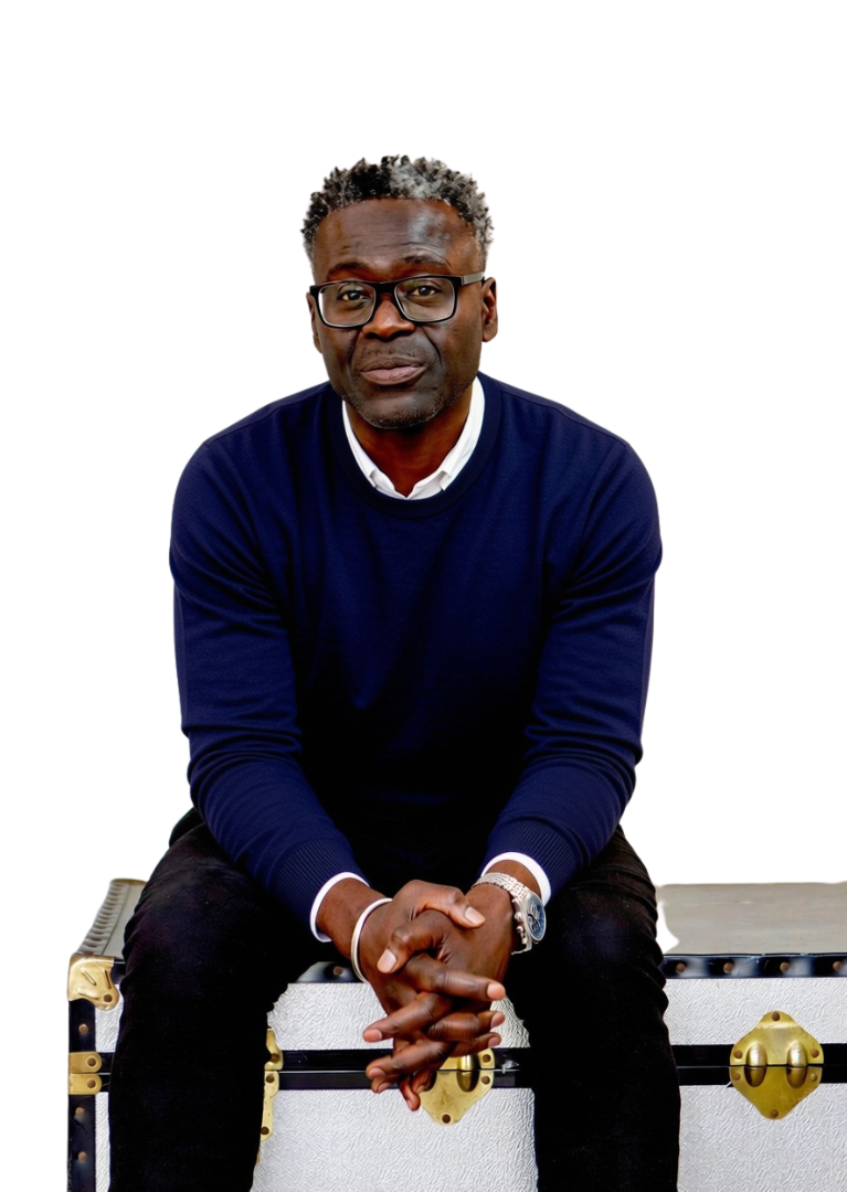 A man with glasses, wearing a navy blue sweater over a white shirt, sitting on a white chest with gold accents against a plain white background.
