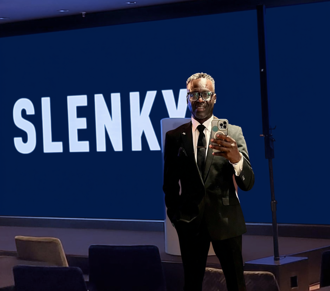 A person in a black suit taking a selfie in front of a presentation screen with the text "SLENKY" on it.