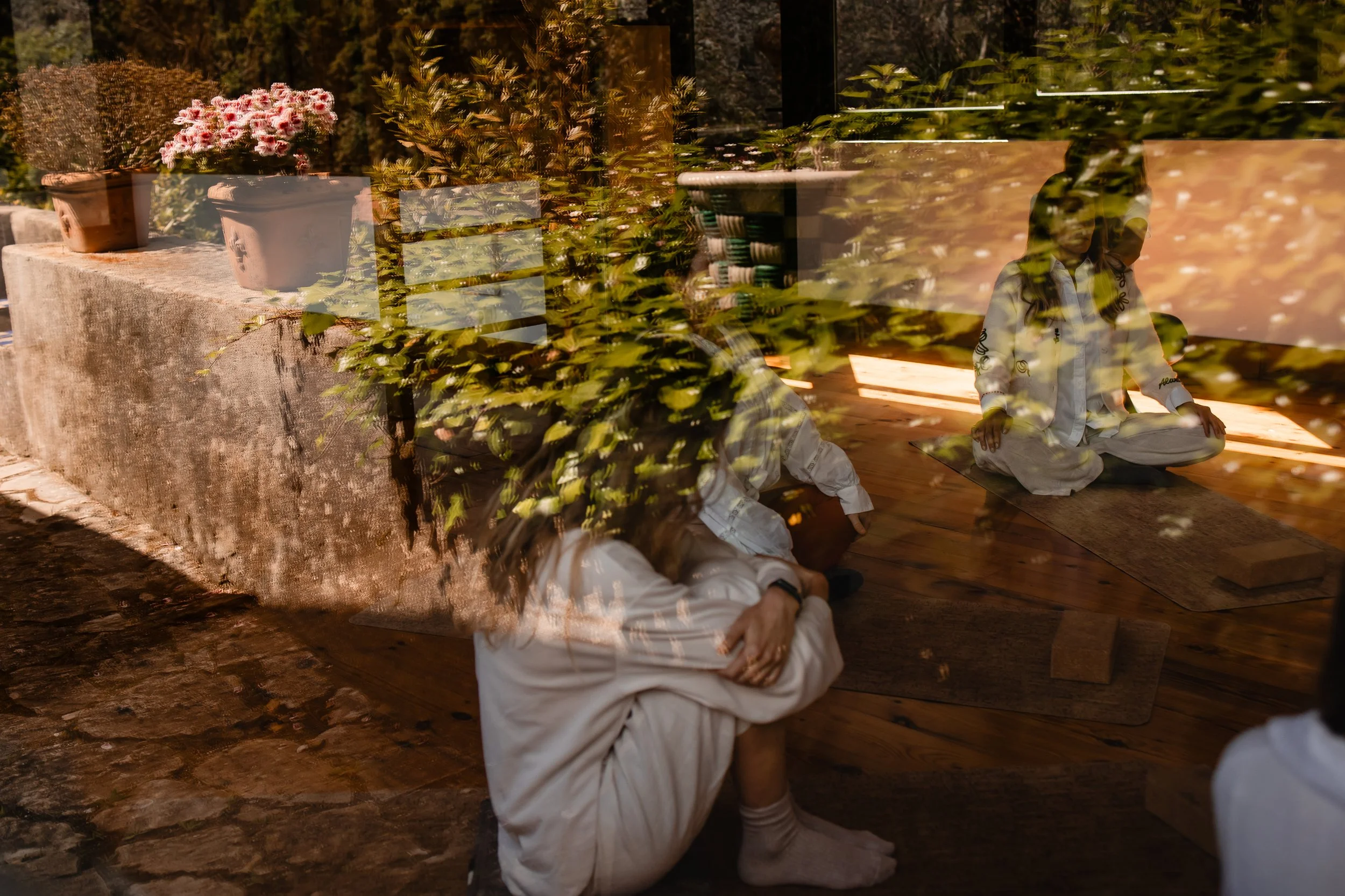 A person dressed in white, sitting cross-legged with hands resting on knees, in a room with wooden flooring. The photo features reflections of potted plants outside on the glass, creating a layered visual effect.